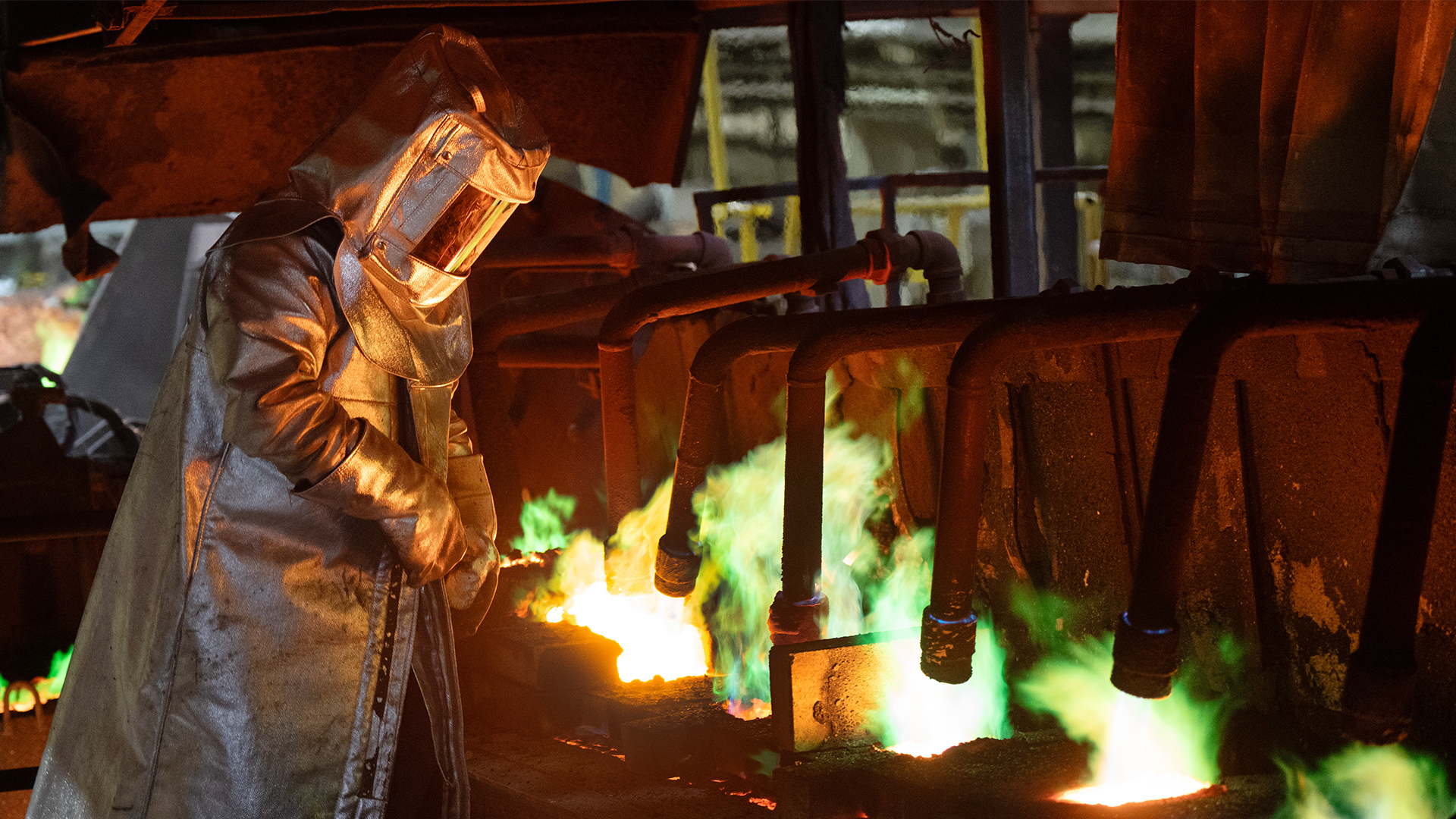 A worker wearing a mask and a silver heat-reflective cloak works near bright green flames.