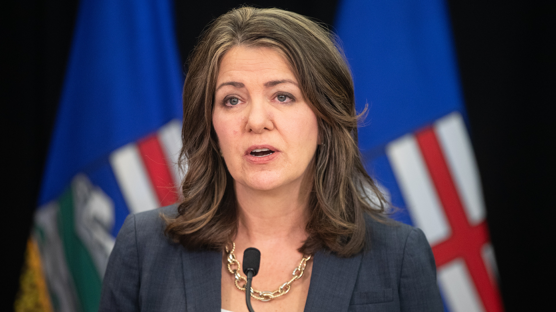 Danielle Smith speaks before microphones with two Alberta flags in the blurred background.