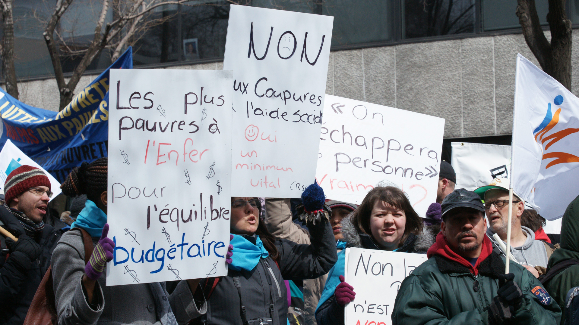 De nombreux individus participent à une manifestation. Certains tiennent des pancartes sur lesquelles on peut lire : « Les plus pauvres à l’Enfer pour l’équilibre budgétaire », « Non aux coupures à l’aide sociale, Oui au minimum vital » et « On n’échappera personne. Vraiment? »