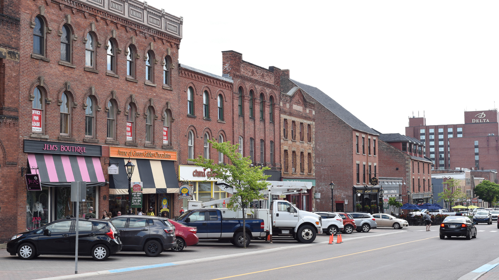 Cars park on the diagonal in front of a row of ornate brick shops that have interesting window patterns, some with curves at the top. A couple of the shops have striped awnings.