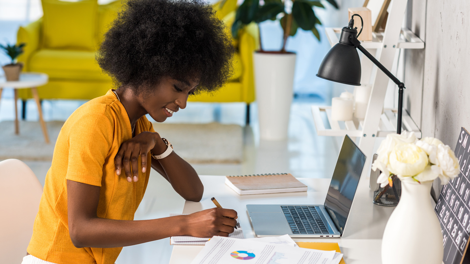 A smiling young Black woman sits at a white desk with a white chair. In front of her is a laptop, a notebook, a couple pages of paper with charts. She is writing in a notebook. A yellow sofa and a white rug, a small three-legged side table with a small potted plant and a larger houseplant are behind her.