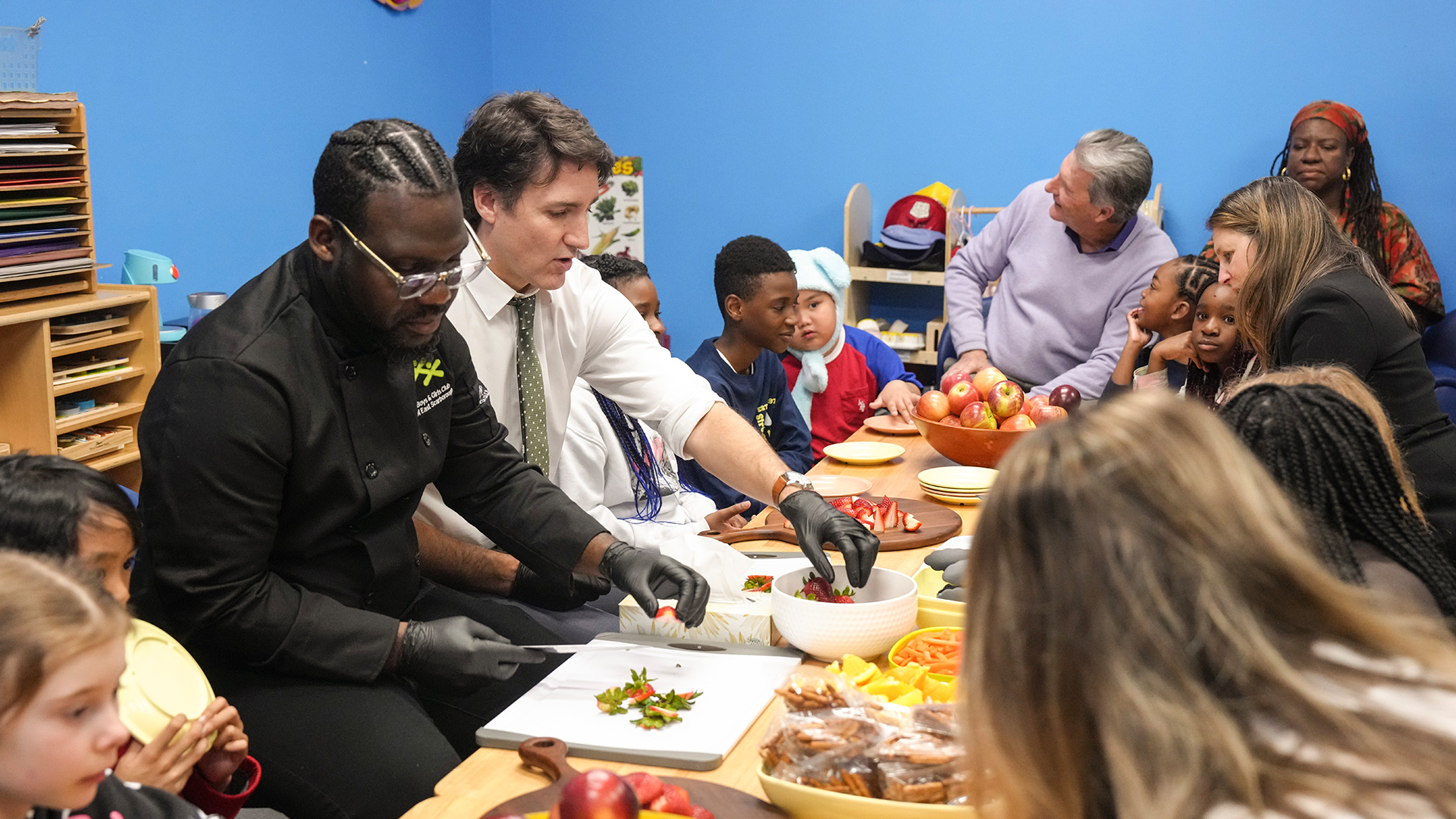 The prime minister has the sleeves of his white dress shirt rolled up. He’s wearing black gloves while reaching for strawberries from a bowl. Several children seem bemused.