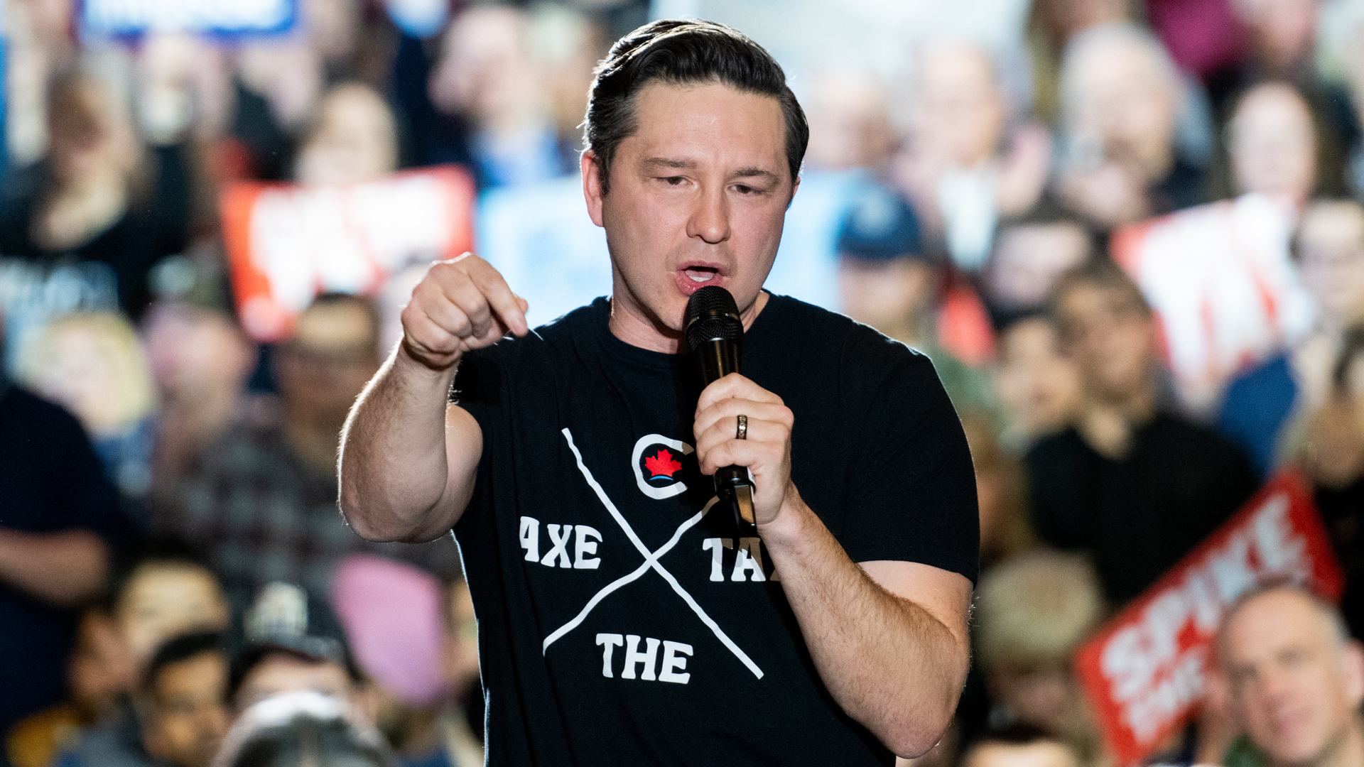 Pierre Poilievre on stage speaking into a microphone wearing a black T-shirt with the slogan “Axe the Tax.”