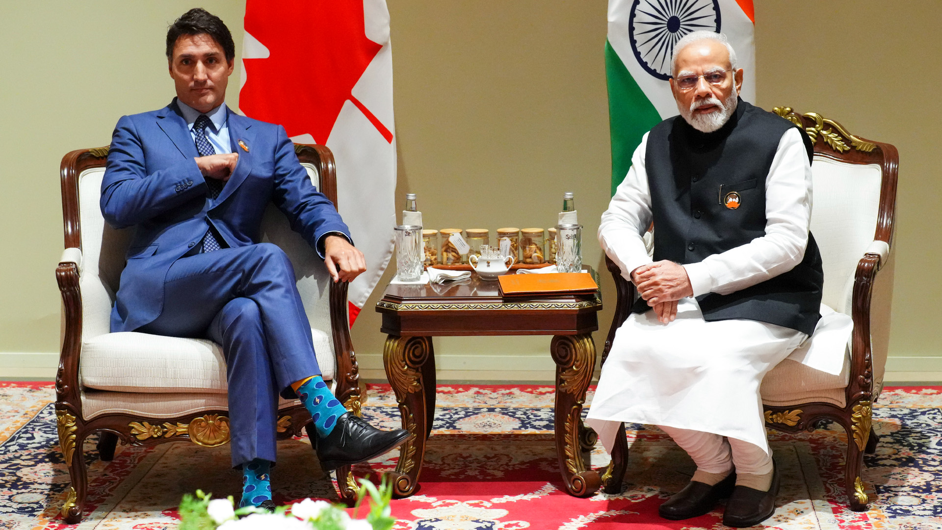 Vêtu d’un complet bleu, le premier ministre canadien Justin Trudeau est assis bien droit dans une chaise. À côté de lui, le premier ministre indien Narendra Modi est également assis sur une chaise, les mains liées, et regarde à l’extérieur du champ de la caméra.