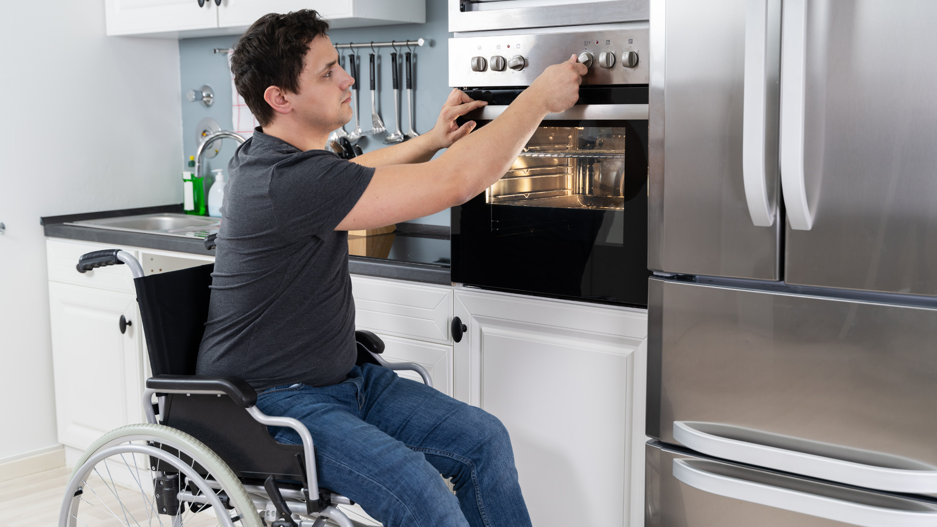 A young man in a wheelchair works in his adapted kitchen. He regulates the temperature of his stainless steel oven.