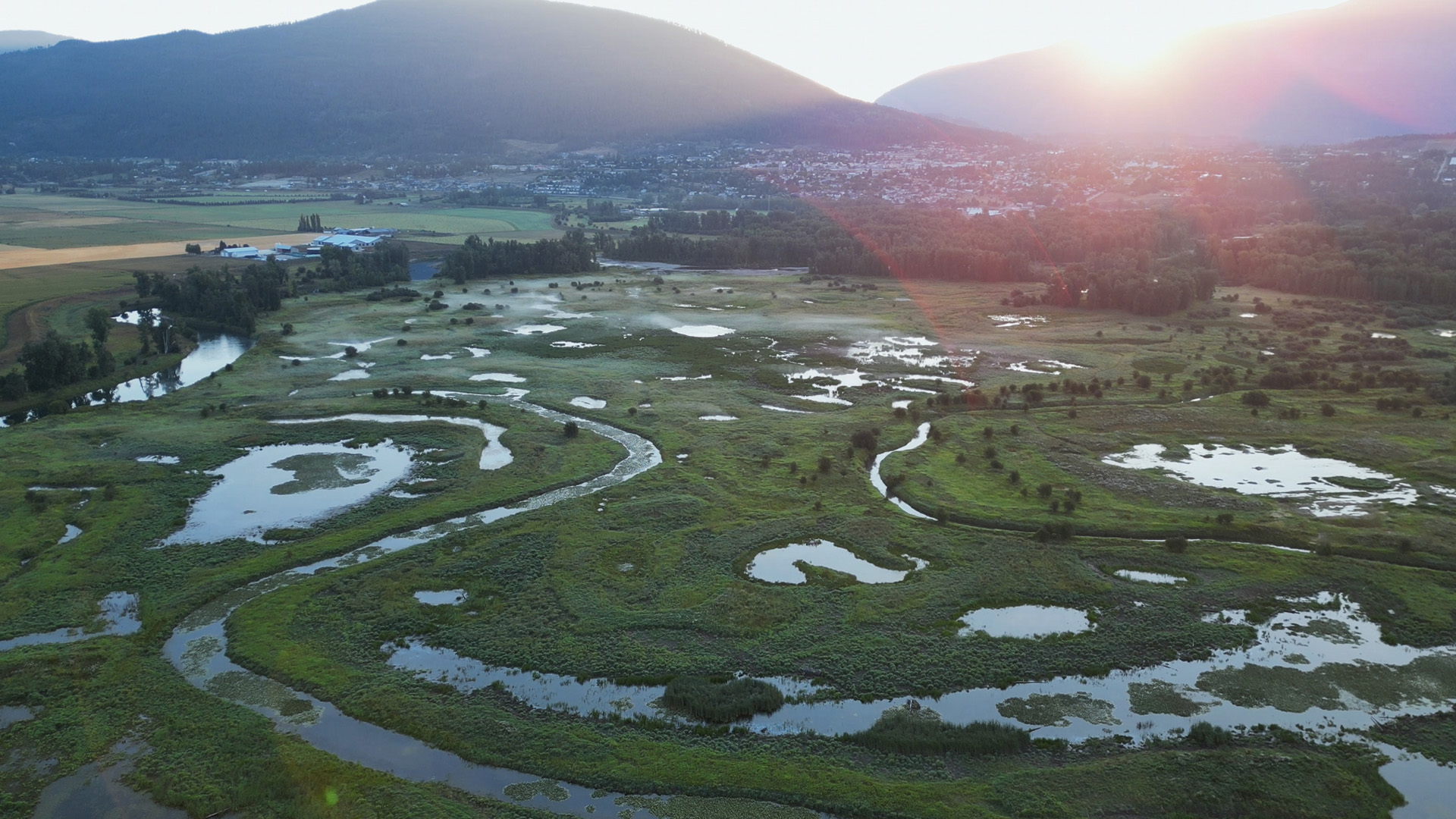 A small river snakes through a lush landscape with several ponds and swales, with a town, agricultural land, and mountains in the background.
