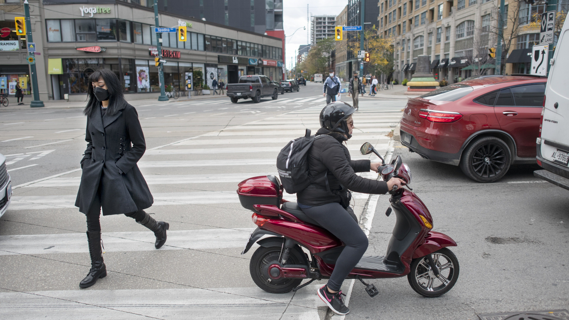 It’s a wide intersection, with three pedestrians in the crosswalk, and a person on a motor scooter in traffic along with cars and trucks.