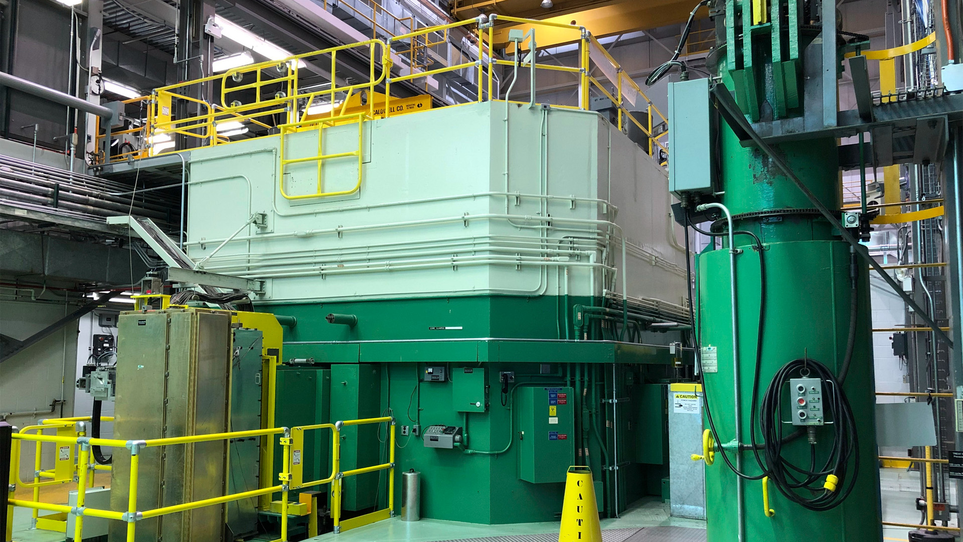 A non-descript steel box painted in two tones of green that appears to be about the size of an extra-tall two-car garage with a yellow ‘caution’ cone in front of it.