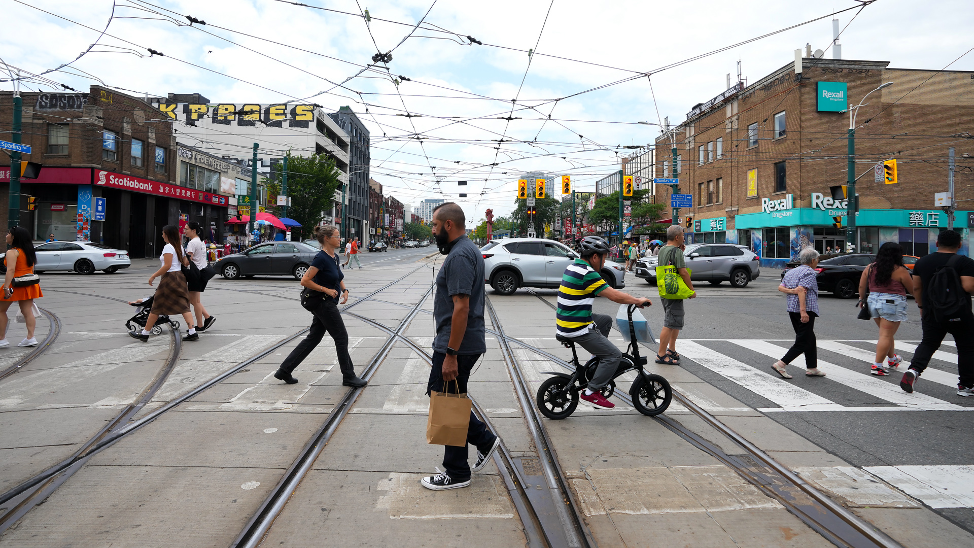 A steady stream of people cross in both directions, walking over tram lines and under a web of electric lines that power the streetcars.
