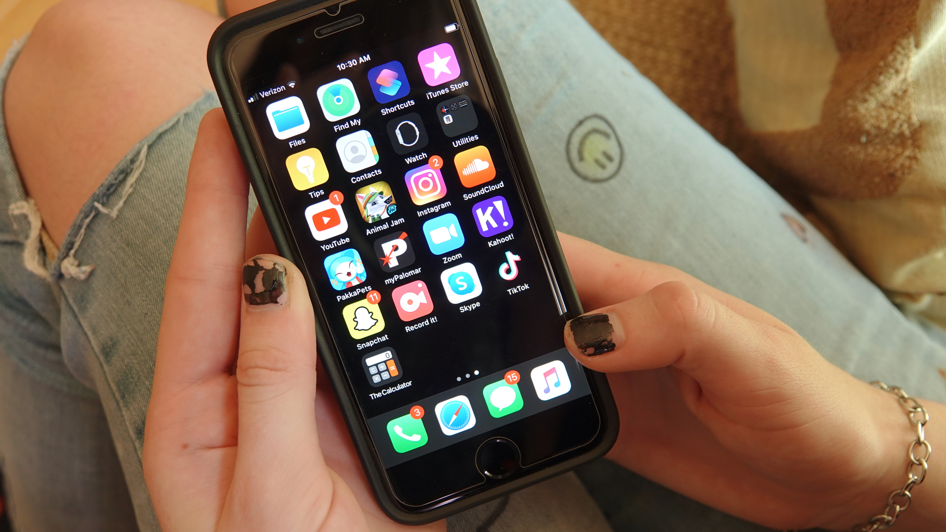 Closeup of an iphone with a home screen full of social-media apps. The person holding it has dark brown chipped nail polish and faded jeans with a smiley face drawn on the denim, along with a large rip in the knee.