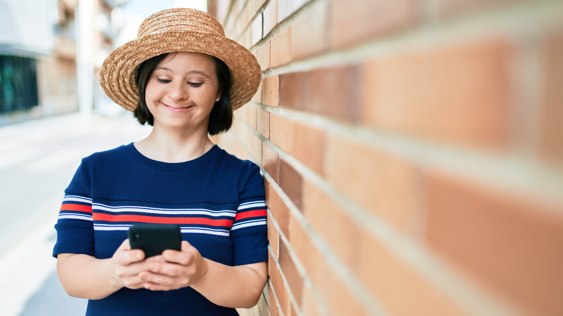 Young woman with down syndrome leans against a brick wall while she texts on a cell phone. She has a straw hat and a navy blue short-sleeved shirt with white and red stripes across it.