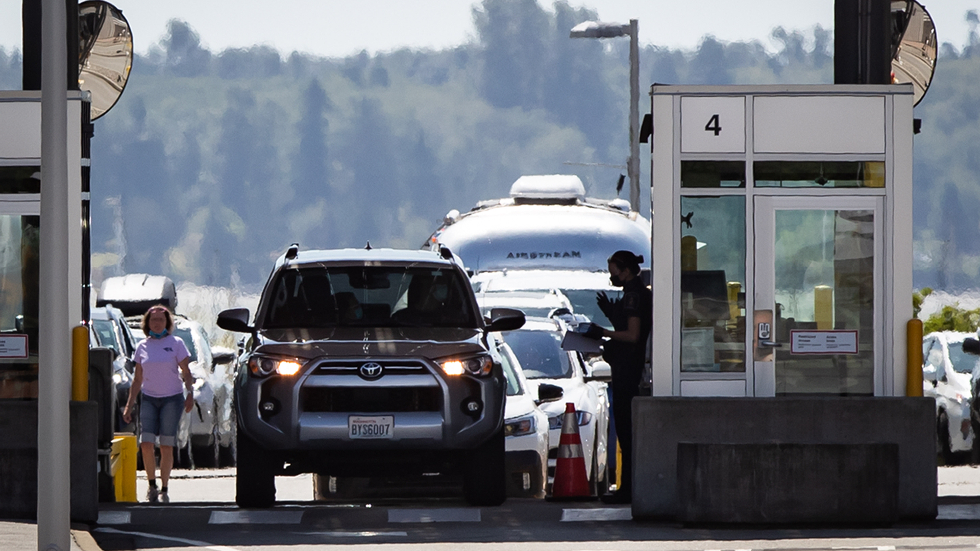 A person in a uniform standing in front of their booth speaks to a driver in a silver Toyota SUV. Several vehicles are lined up behind them.