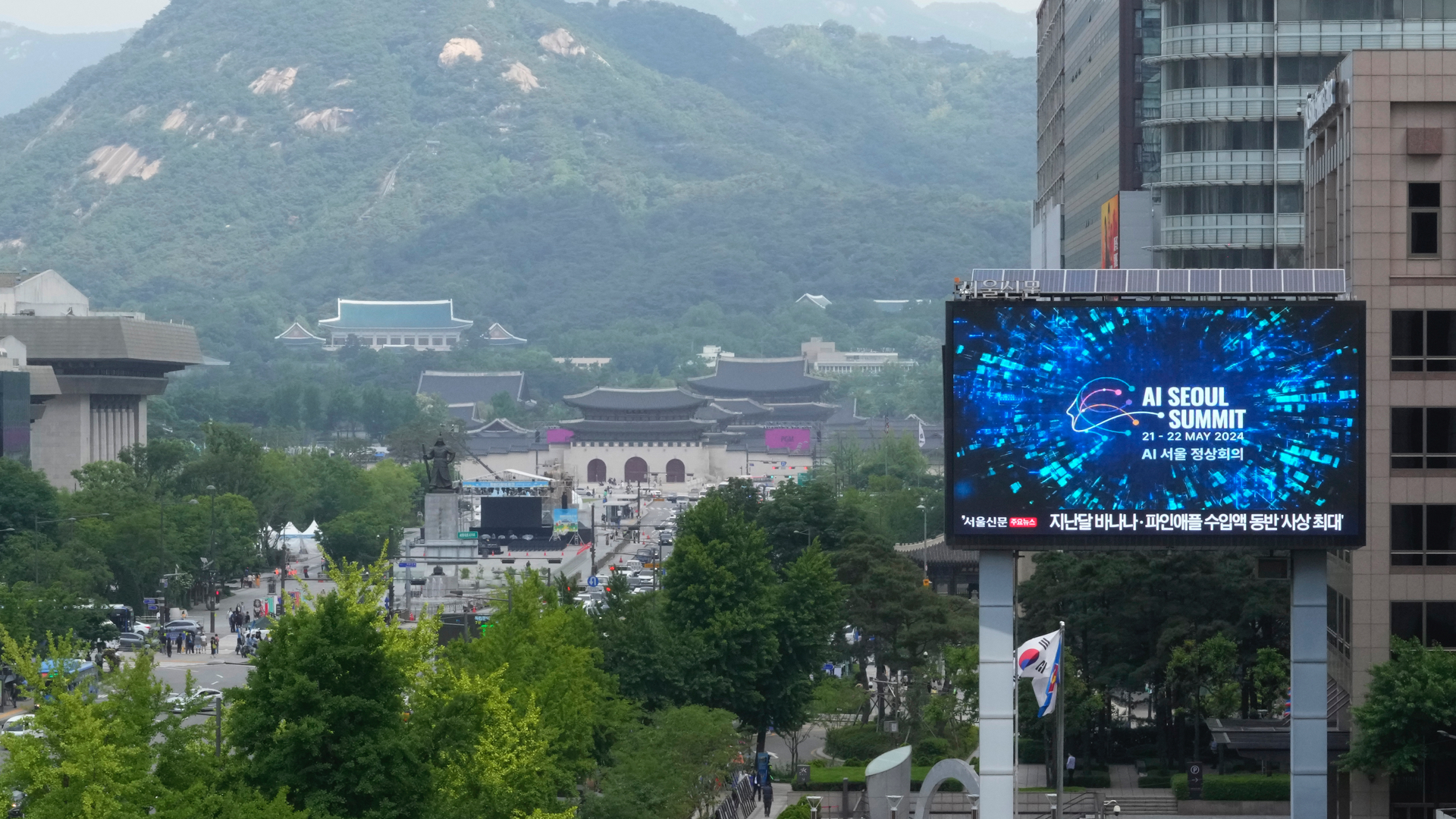 The screen is a large billboard sitting high above the ground next to a few tall buildings along a tree-lined street. In the distance are two temples with curved roofs with a green-covered mountain behind them.