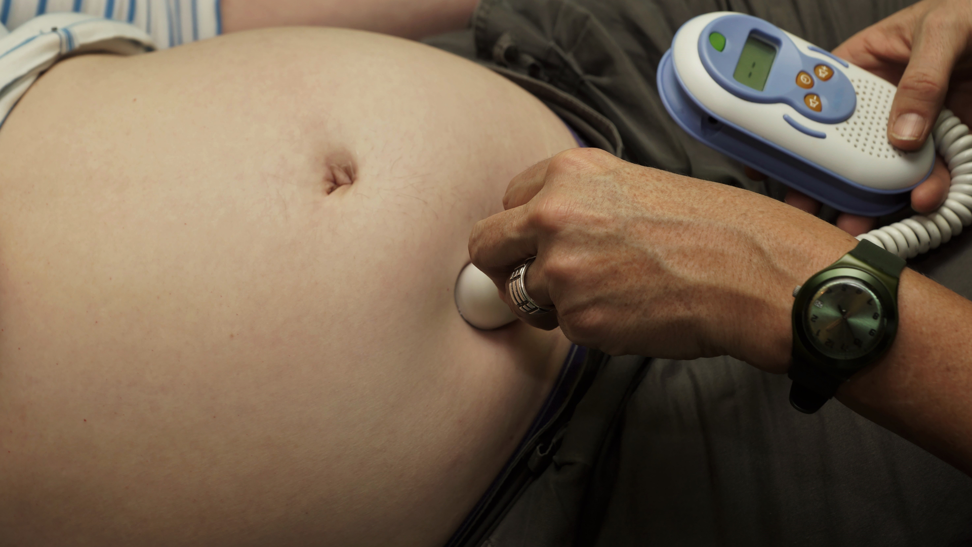 Tight crop around a woman’s bare belly, where a handheld monitor is attached to a small, metal sensor being held to the skin.