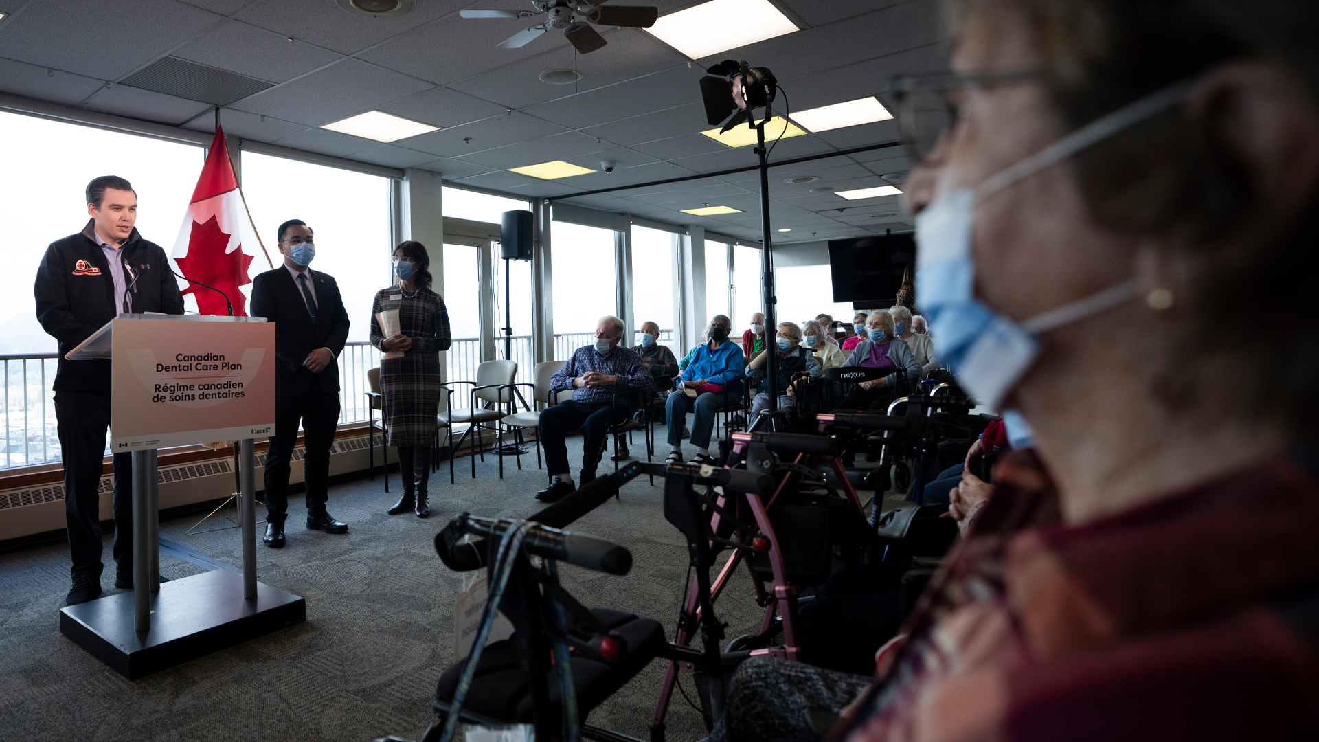 Beech speaks at a podium in front of a wall of windows. A Canadian flag hangs nearby. Several rows of older people are seated as they listen.