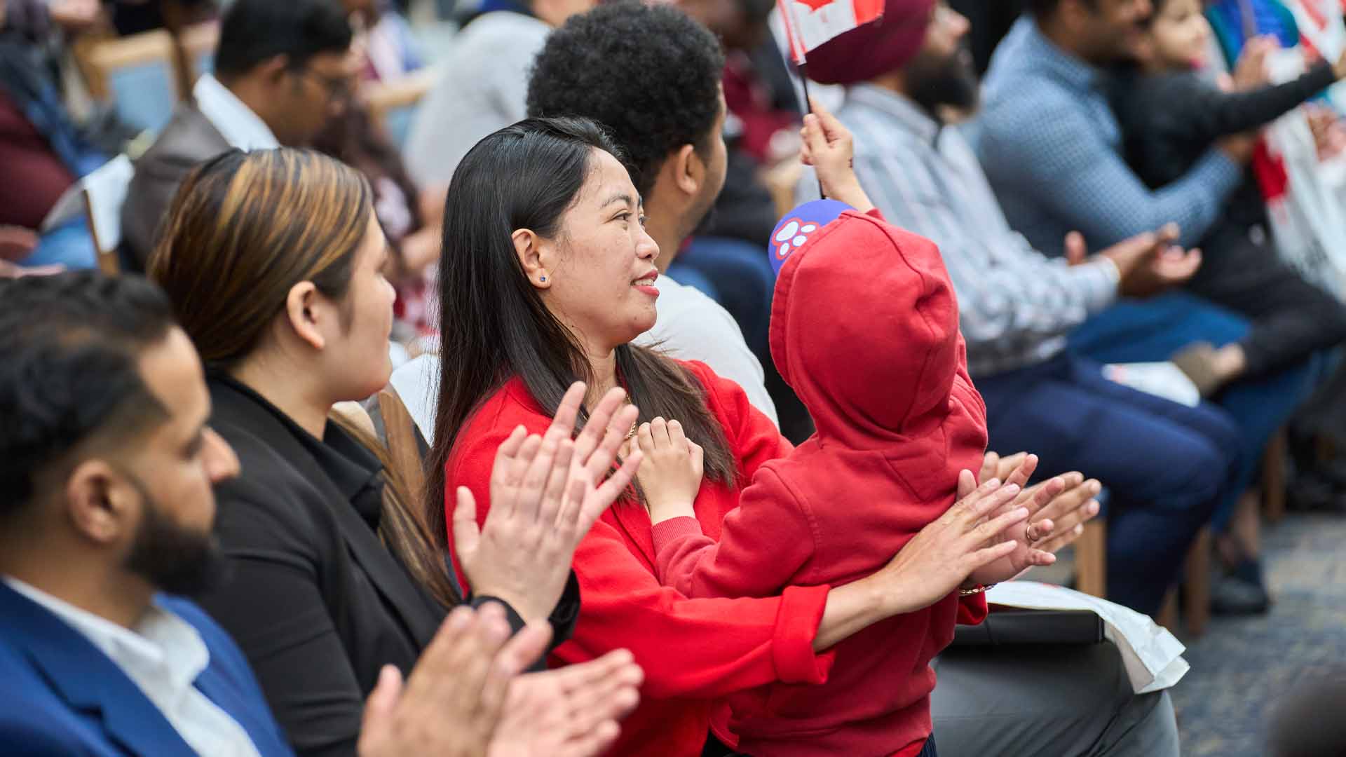 Martin is one of several dozen people pictured sitting in four rows of chairs. Most everyone is clapping. Martin is smiling. Kale holds up a small Canadian flag. He and his mum are dressed in red.