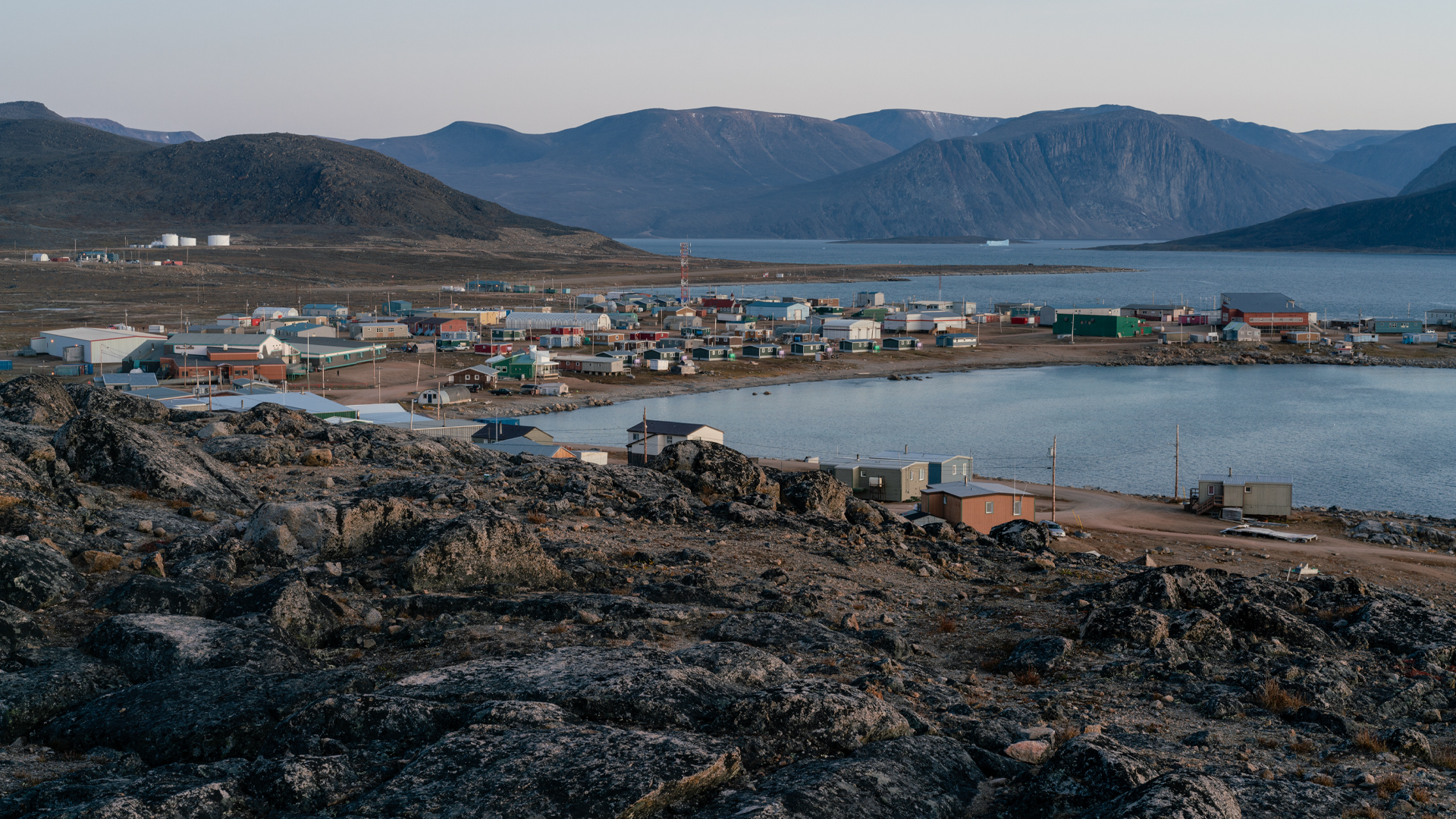 Several dozen one- and two-storey buildings are clustered near a body of water with mountains in the near distance. Dark rocks sit in the foreground. There are no trees.