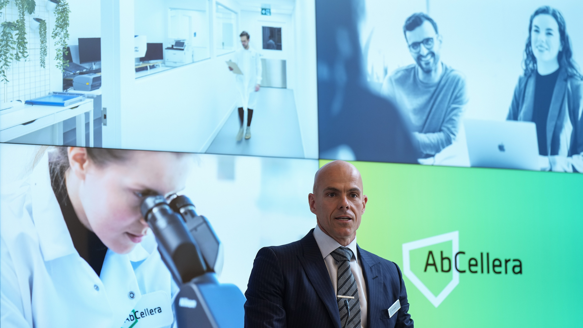 Hansen stands in front of a large screen split into six images depicting some of the people in the company. One person looks into a microscope. Two others sit in front of a laptop. A third walks down a hallway, clipboard in hand.