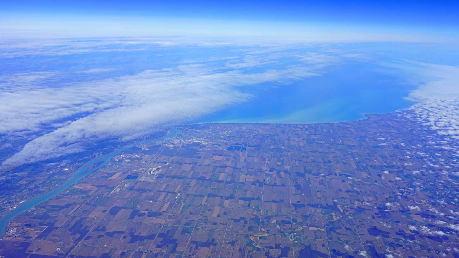 A few thin clouds sit above an expanse of blue in the distance. In the foreground is a land mass that is made of squares of brown, green and dark blue.