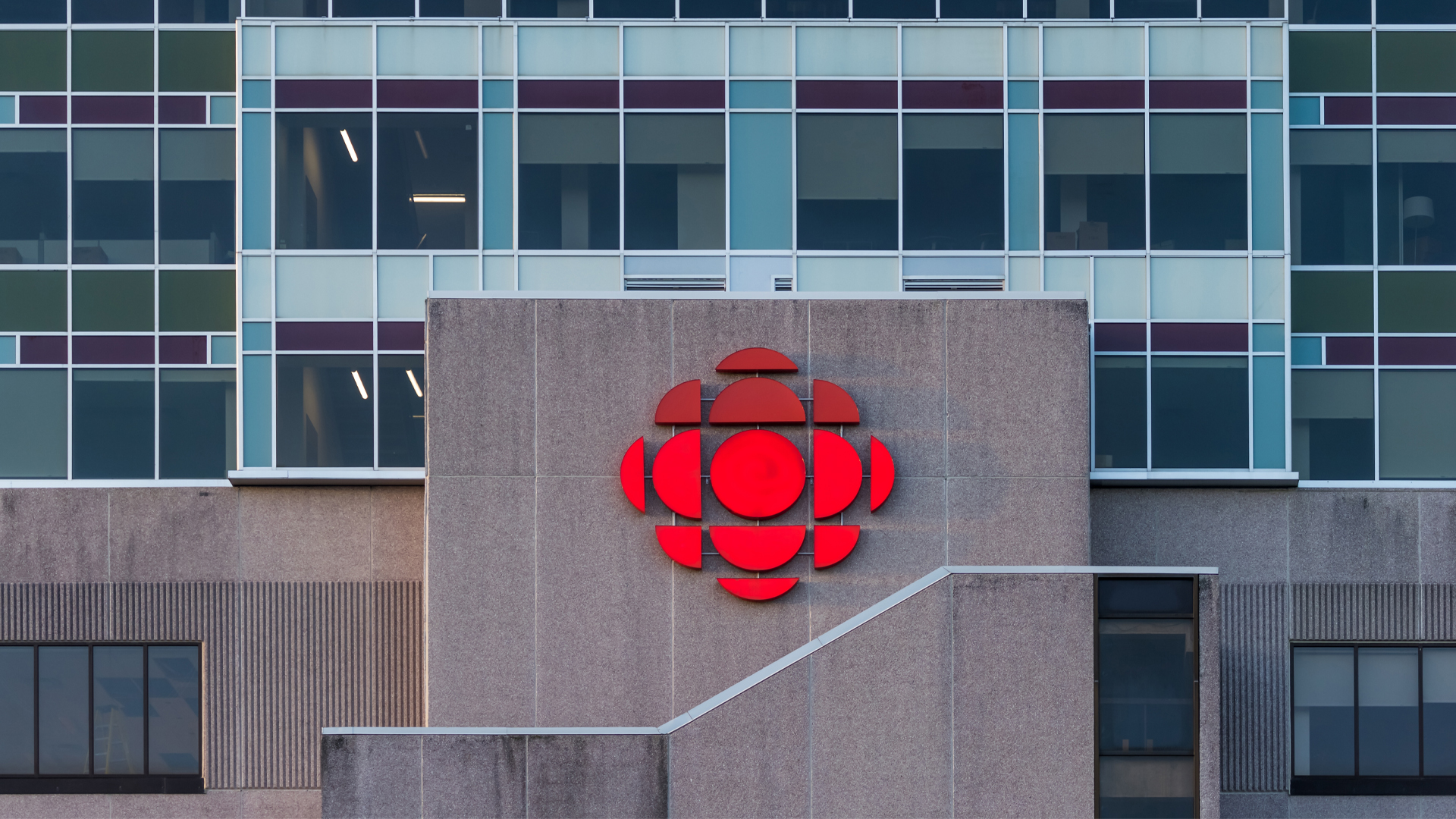The red CBC symbol, a circle with two layers of half-moons surrounding it, adorns the stone-wall entrance. Behind it is a wall of windows with colour panels in shades of turquoise, burgundy and pale green. The window panes are tinted.