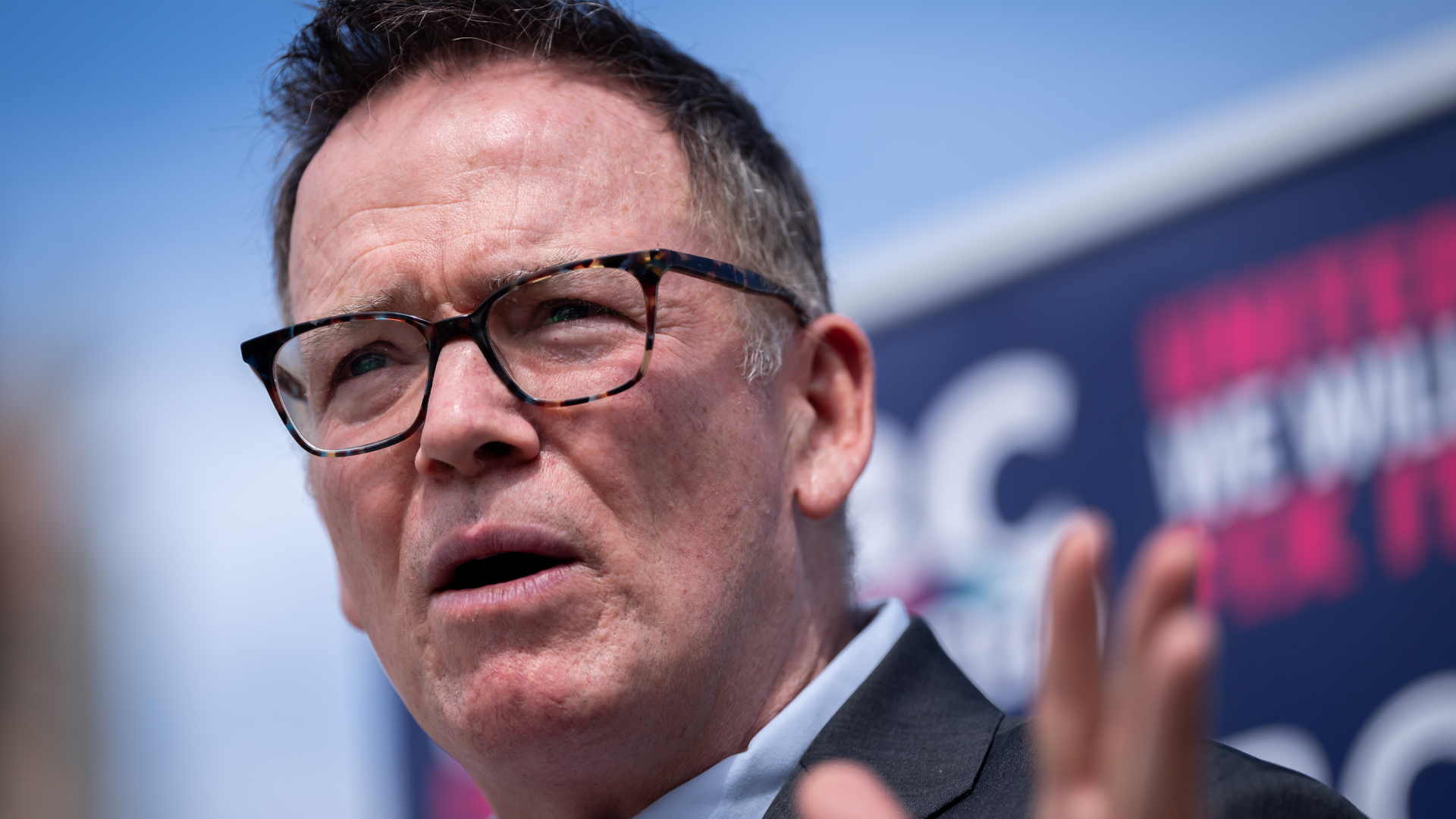 A head-and-shoulders shot of him. He is wearing brown eyeglasses, a dark grey jacket and light blue shirt. His hair is dark and short, with grey at the temples.