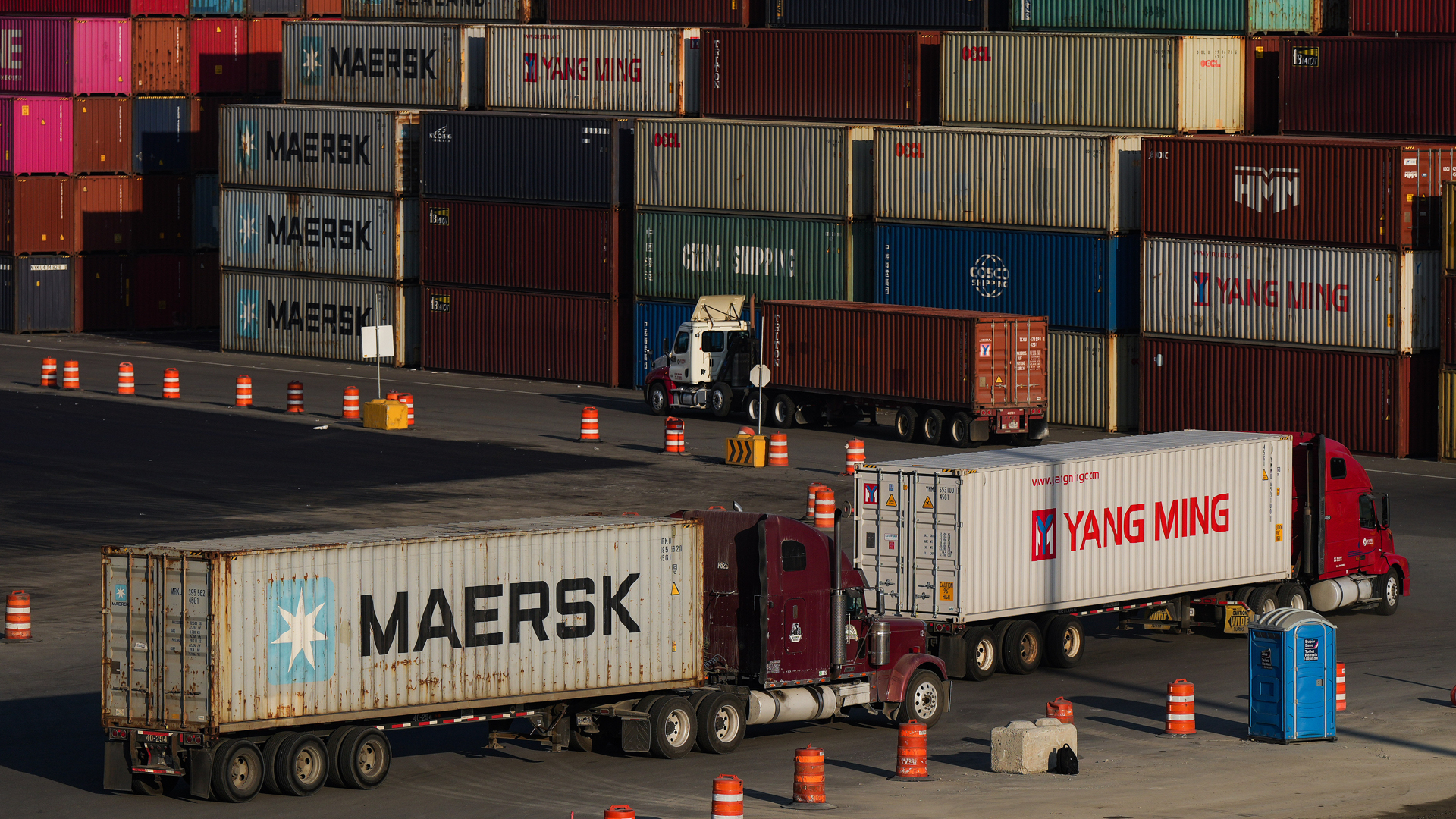 Three trucks with cargo containers drive into an area with stacks of containers.