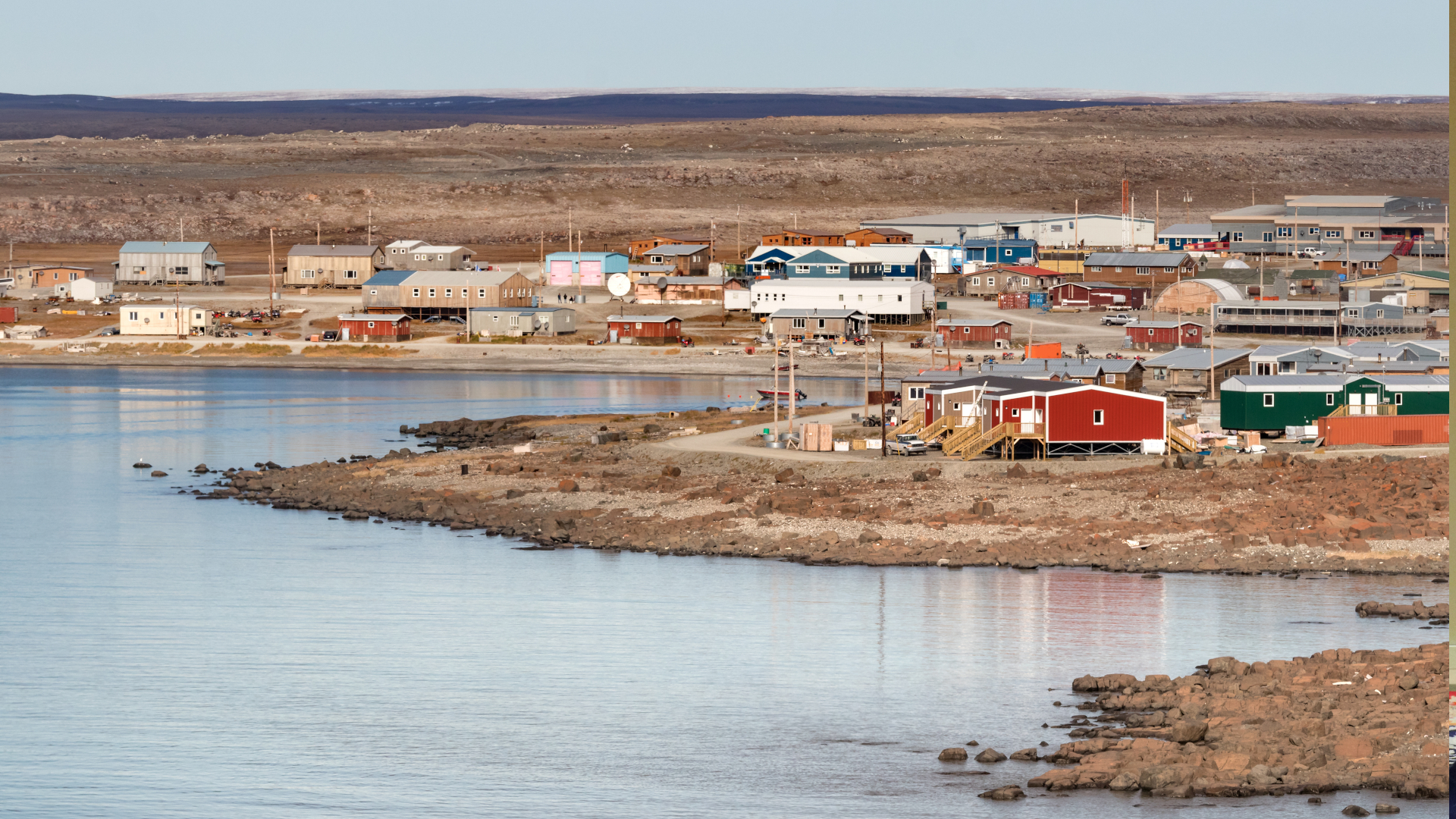 On a rocky coastline, a small cluster of bungalows and similar buildings sit. In the distance, the land is flat and brown.