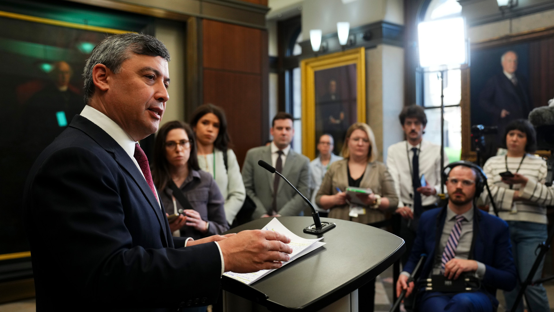 Chong stands at a podium in front of a nearly a dozen reporters. On the walls are large framed portraits of men. The walls are panelled in wood, the windows are tall and arched at the top.