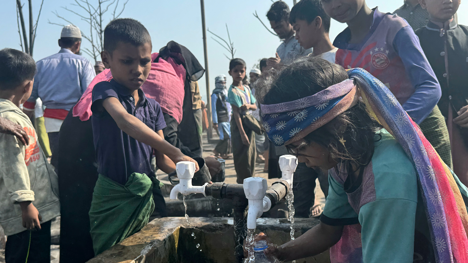 A child wearing a scarf around their head fills a plastic water bottle at a communal tap.