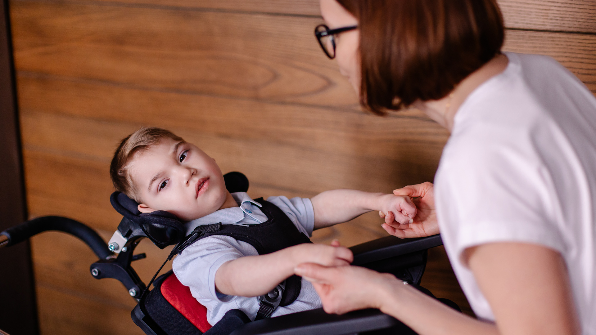 A child strapped into a wheel chair inclined slightly backwards holds hands with his mother who is talking to him.