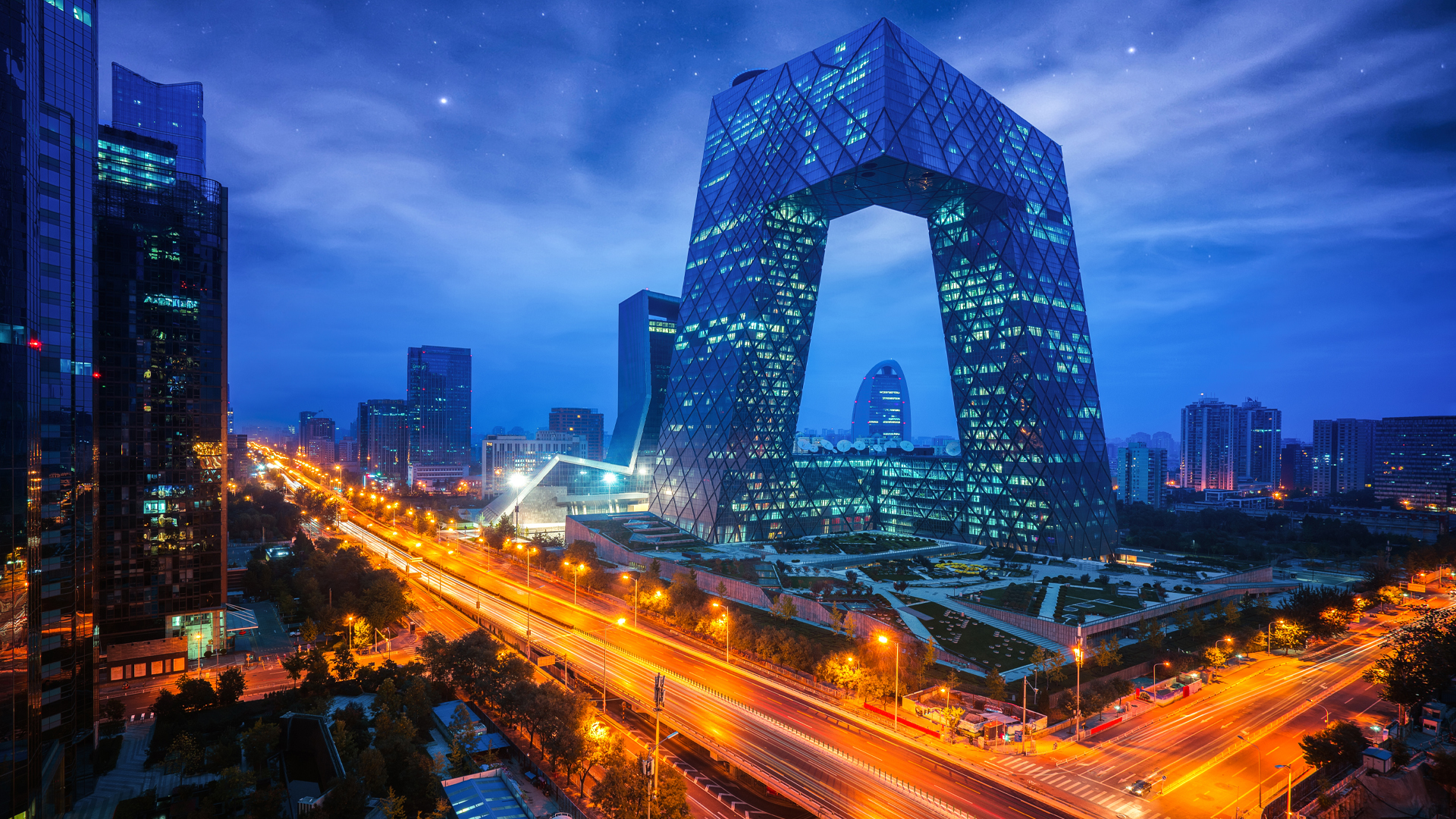 Contemporary architecture is showcased in this photo of the city at dusk, where the headlights of passing cars leave golden streaks. Skyscrapers frame the skyline. In the middle are two towers connected at the top and bottom.
