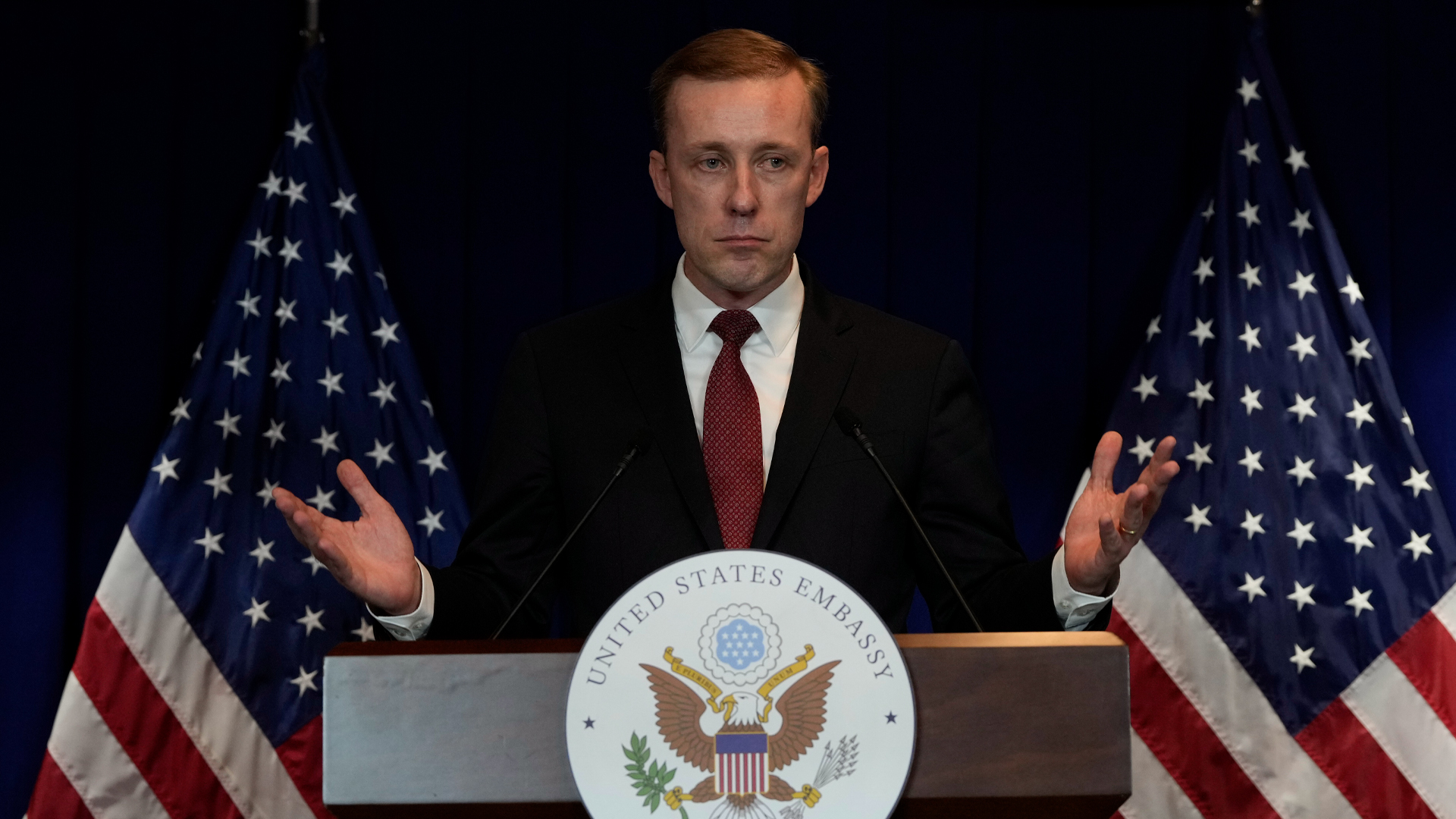 Sullivan stands at a podium that has a round white crest that says United States Embassy Beijing. It is illustrated with an eagle with wide brown wings, and the bird is holding a leaf and a handful of arrows. Two American flags stand draped behind Sullivan, who is dressed in a dark suit and isn’t smiling.