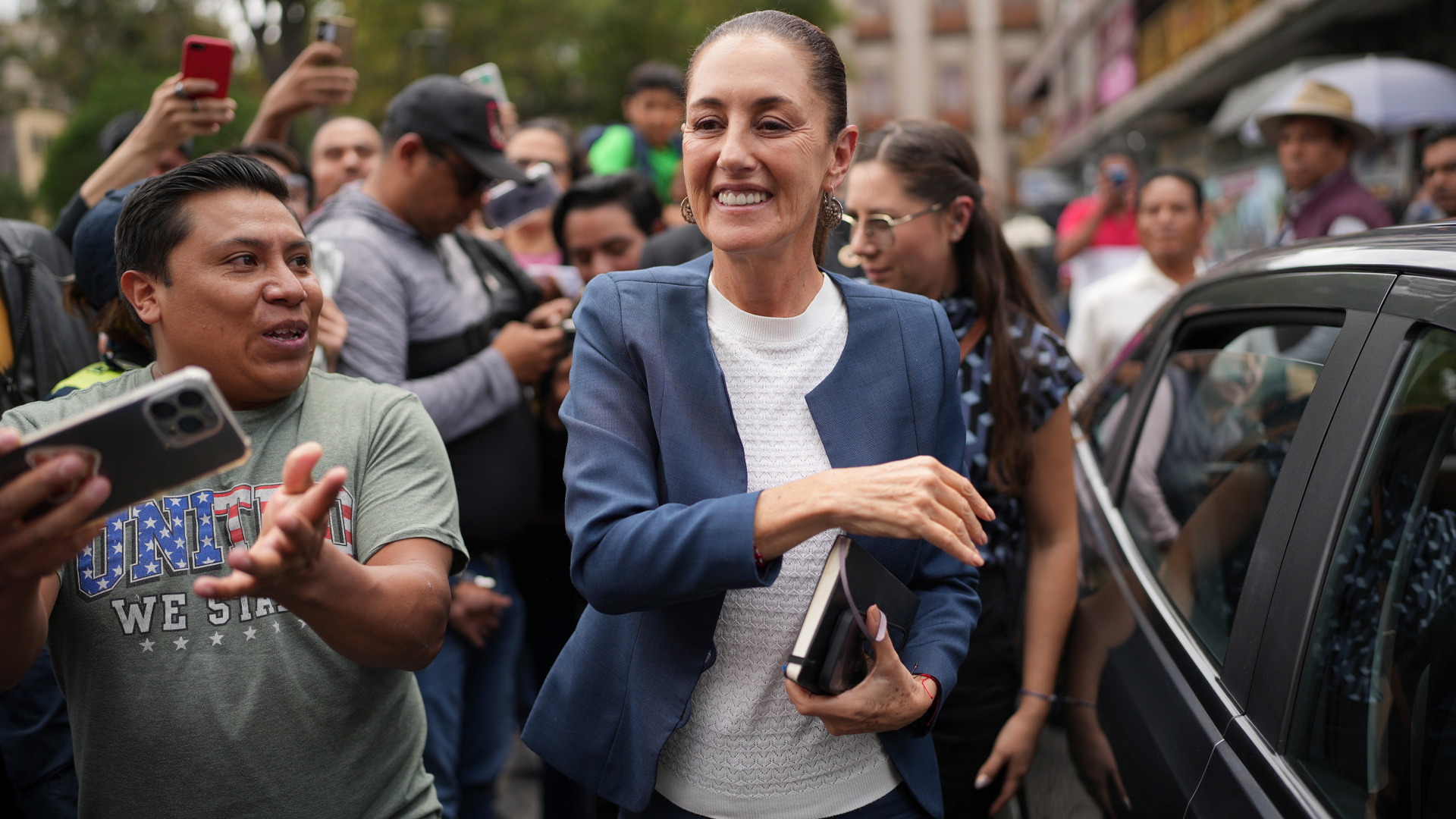 Sheinbaum is walking next to a black car and is surrounded by men and women. She is smiling. A man next to her is in a T-shirt that says “United we stand.” Some people are taking pictures with their phones, holding them up.