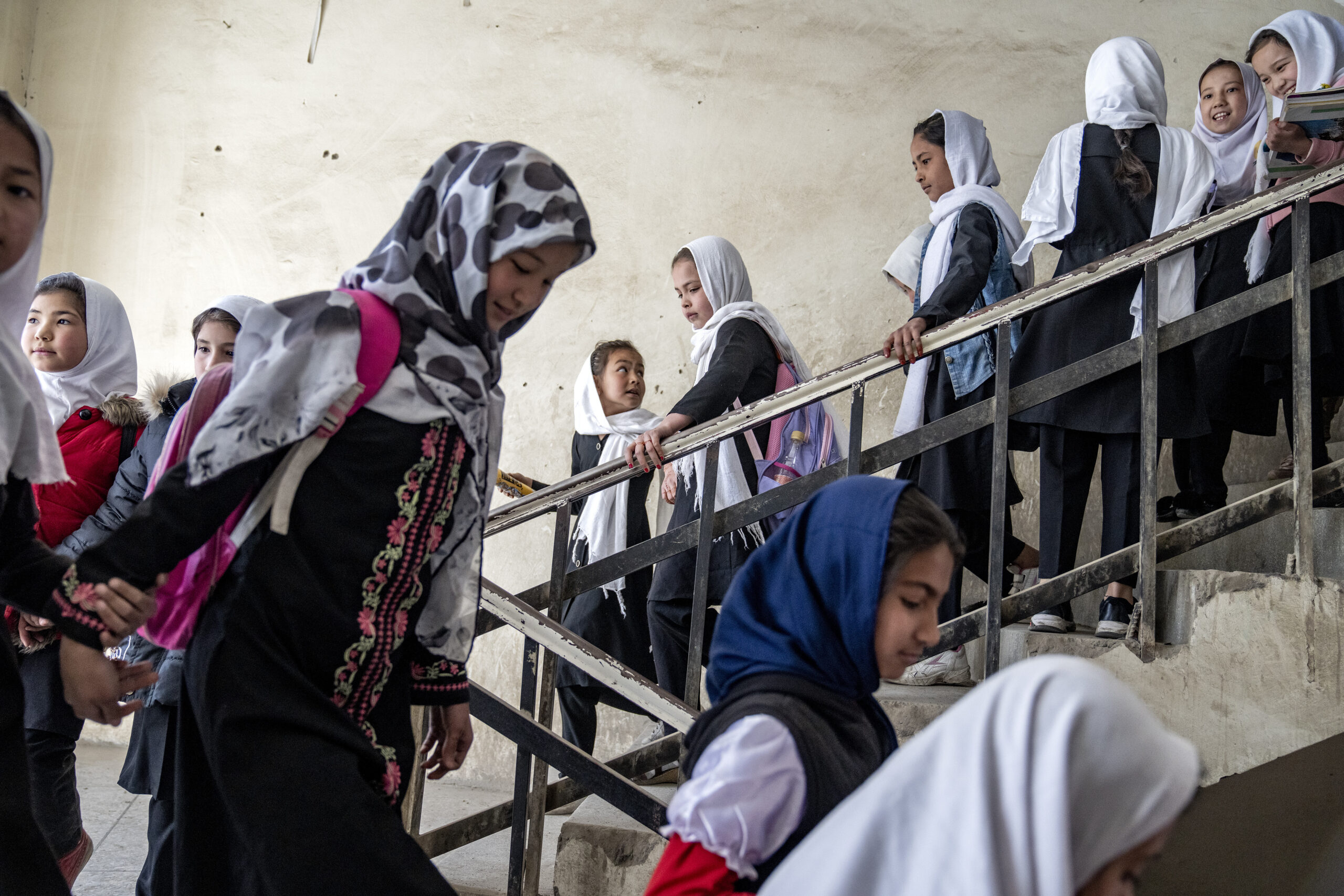 A set of stairs is filled with girls on their way down. They are all mostly dressed in black, with tunics and pants, and head scarves that are mostly white.
