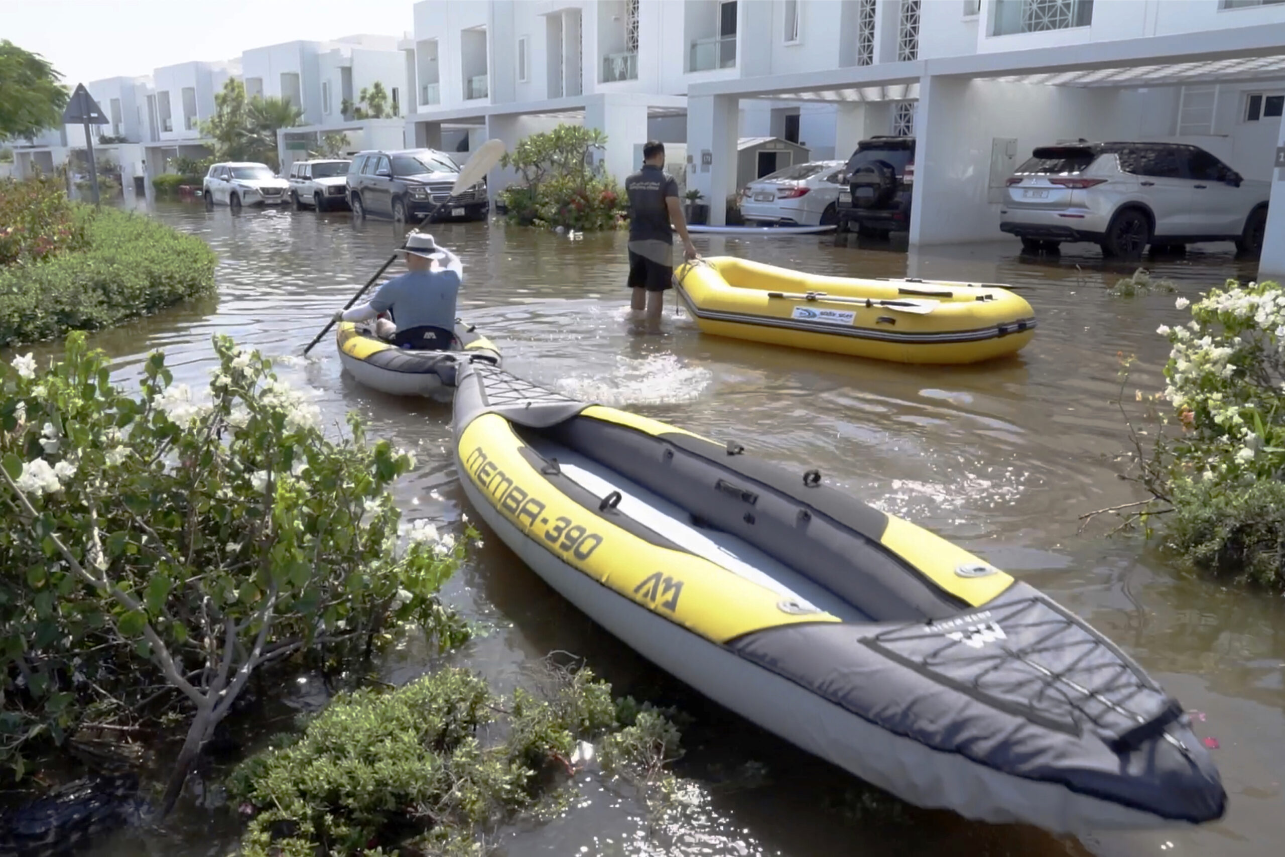 In front of a row of white flat-roof townhouses with balconies and parked cars, the water is knee-high. A man is kayaking in an inflatable kayak that is light grey and yellow. He is towing a second kayak. Another man stands nearby holding on to a yellow inflatable dinghy. Landscaped shrubs are partly submerged.
