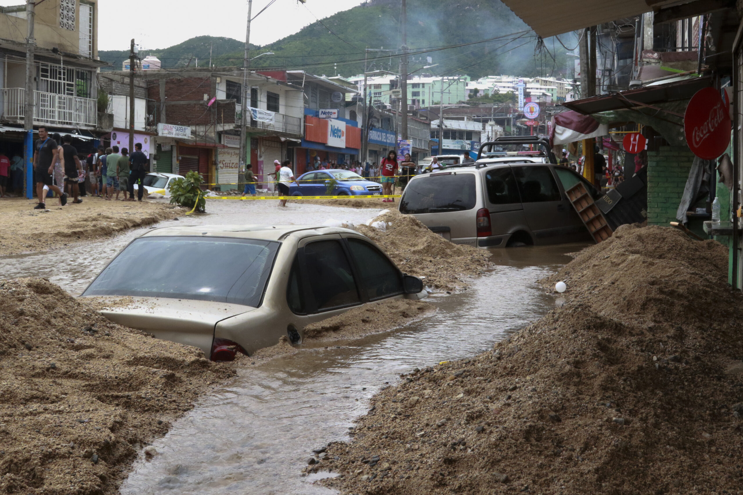 Several cars and trucks are lodged in sand that has piled up around them to the level of the passenger-door windows. The road is filled with two-storey buildings that house shops and apartments. Groups of people walk the road. In the distance is a green-covered hill.