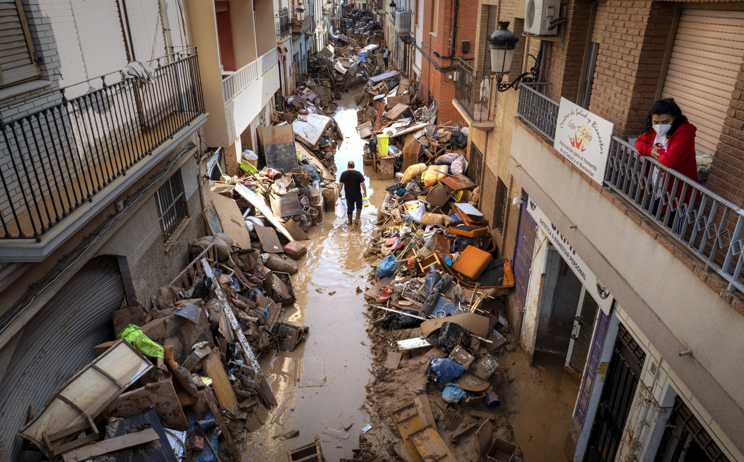 In a narrow street lined by brick and concrete buildings with shops and apartments, a man walks through a small amount of muddy water left from the flood. On both sides of him are tall piles of items all covered in mud. A woman looks down from a second-floor balcony.