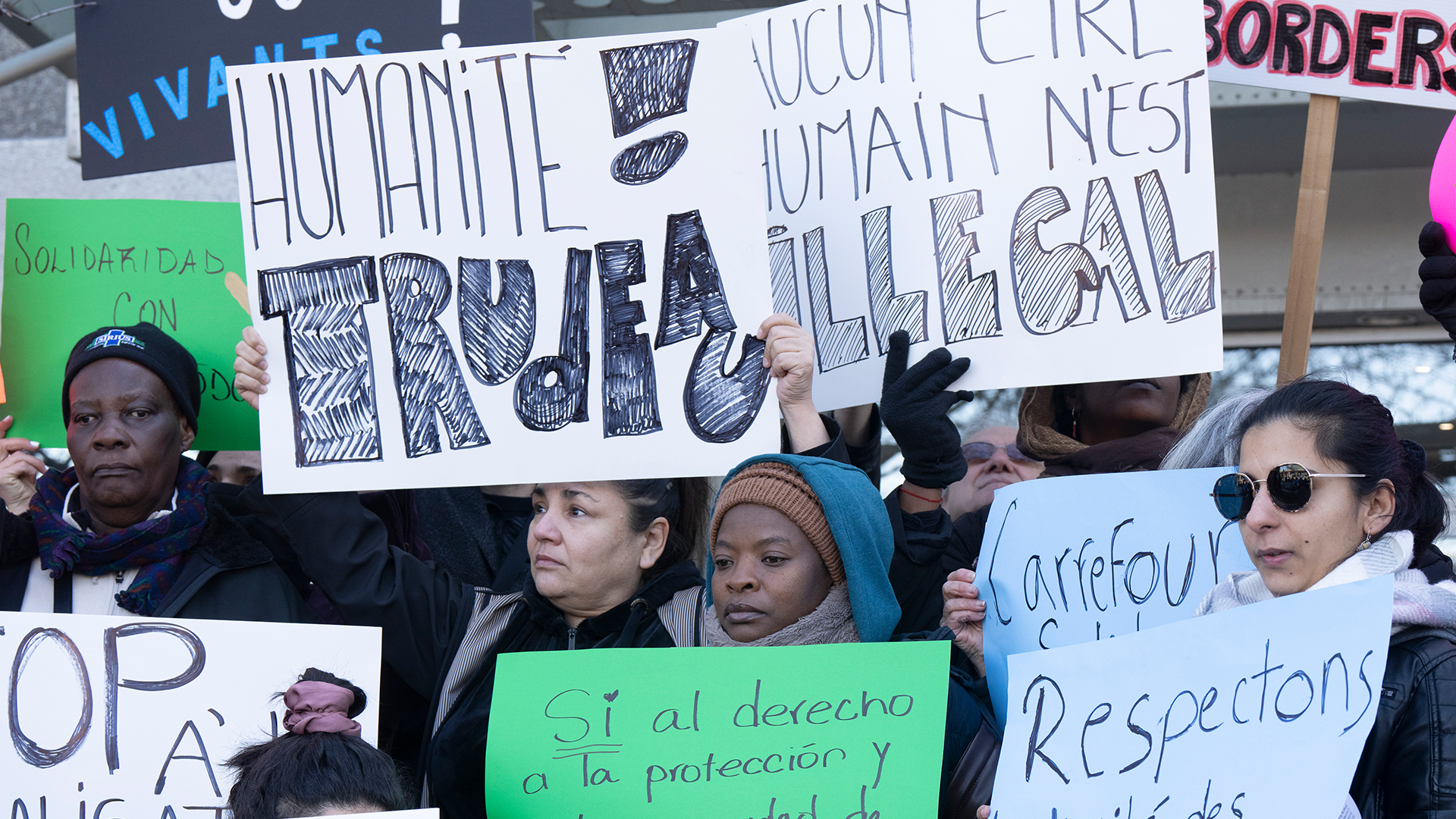 A crowd of people with four protesters in the foreground hold multiingual signs saying “no human is illegal” and “respect the dignity of asylum seekers.”