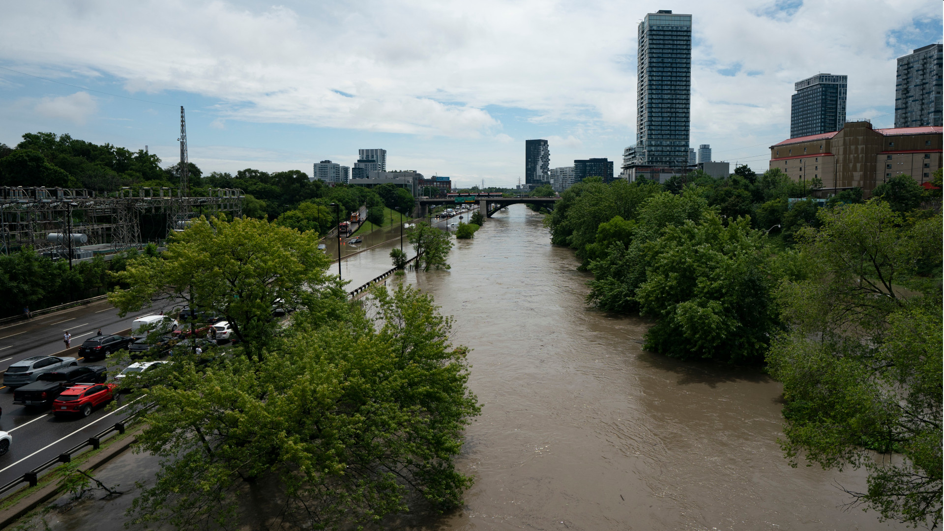 The river runs parallel to the highway. Its waters are brown and moving, spilling onto the lowest stretch of road. Traffic is stopped in several lanes, unable to proceed. The river is lined with some trees. In the distance are some downtown towers.
