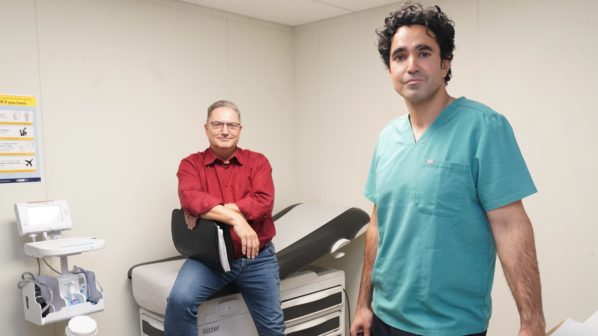 Dr. Boozary on the right is wearing a hospital scrub shirt and is holding a stethoscope. Fred Vitor holds a folder of papers while sitting on an examination table.
