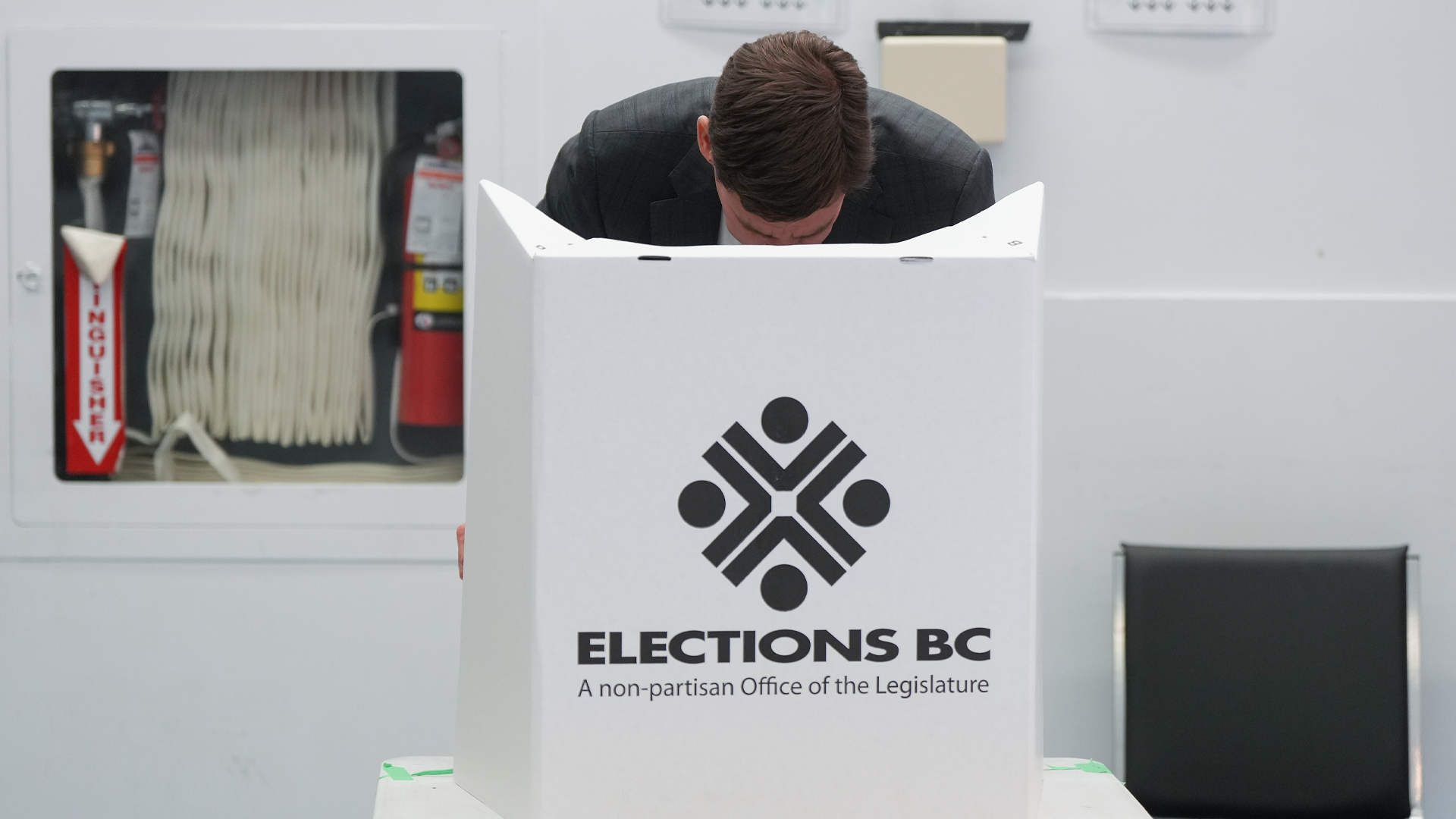 A man crouches behind the white voting screen with “ELECTIONS B.C., a non-partisan office of the legislature” written on it.