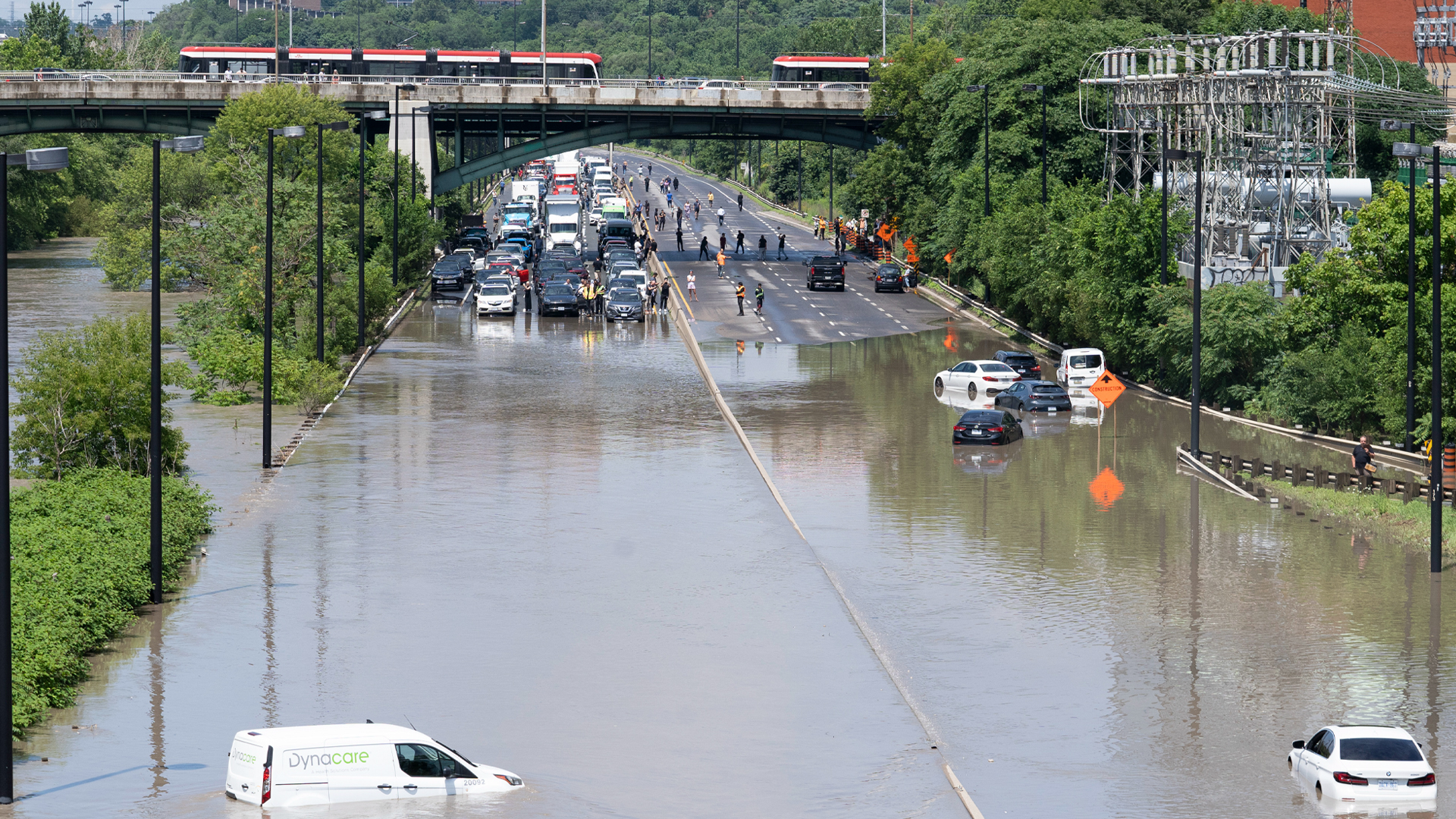 Four lanes of traffic are full with cars siting at a standstill at the edge of what looks like a lake of water that has pooled in a low stretch of the highway. Seven vehicle are partially submerged in the water, which has risen up to near the door handle of a white van in the deepest water, which has streamed in from the banks of the overflowing river that runs next to the road. Thick stands of trees line the road. Two red-and-white TTC trains are visible on an overpass.