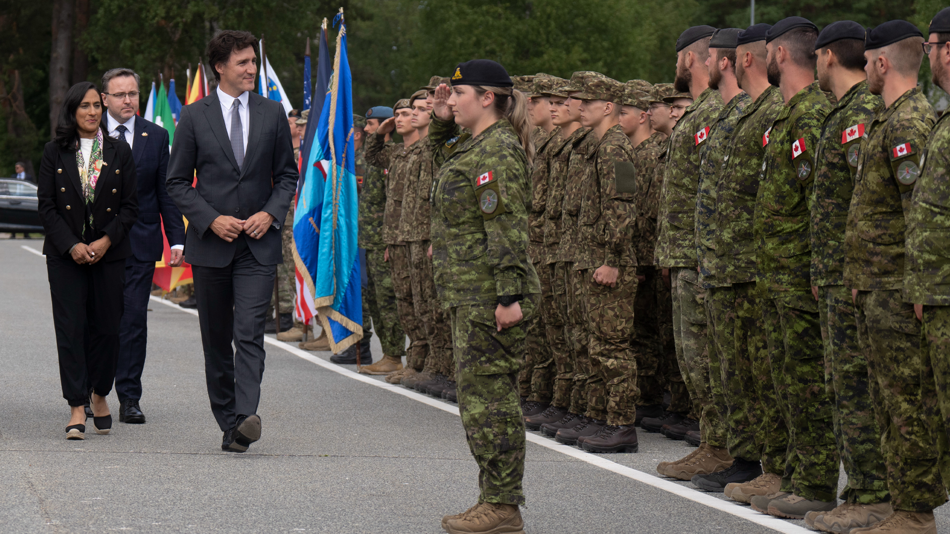 Several dozen soldiers stand shoulder to shoulder in green- and brown-patterned fatigues. Trudeau, Anand and an unidentified man pass look at the group as they pass in front of it.