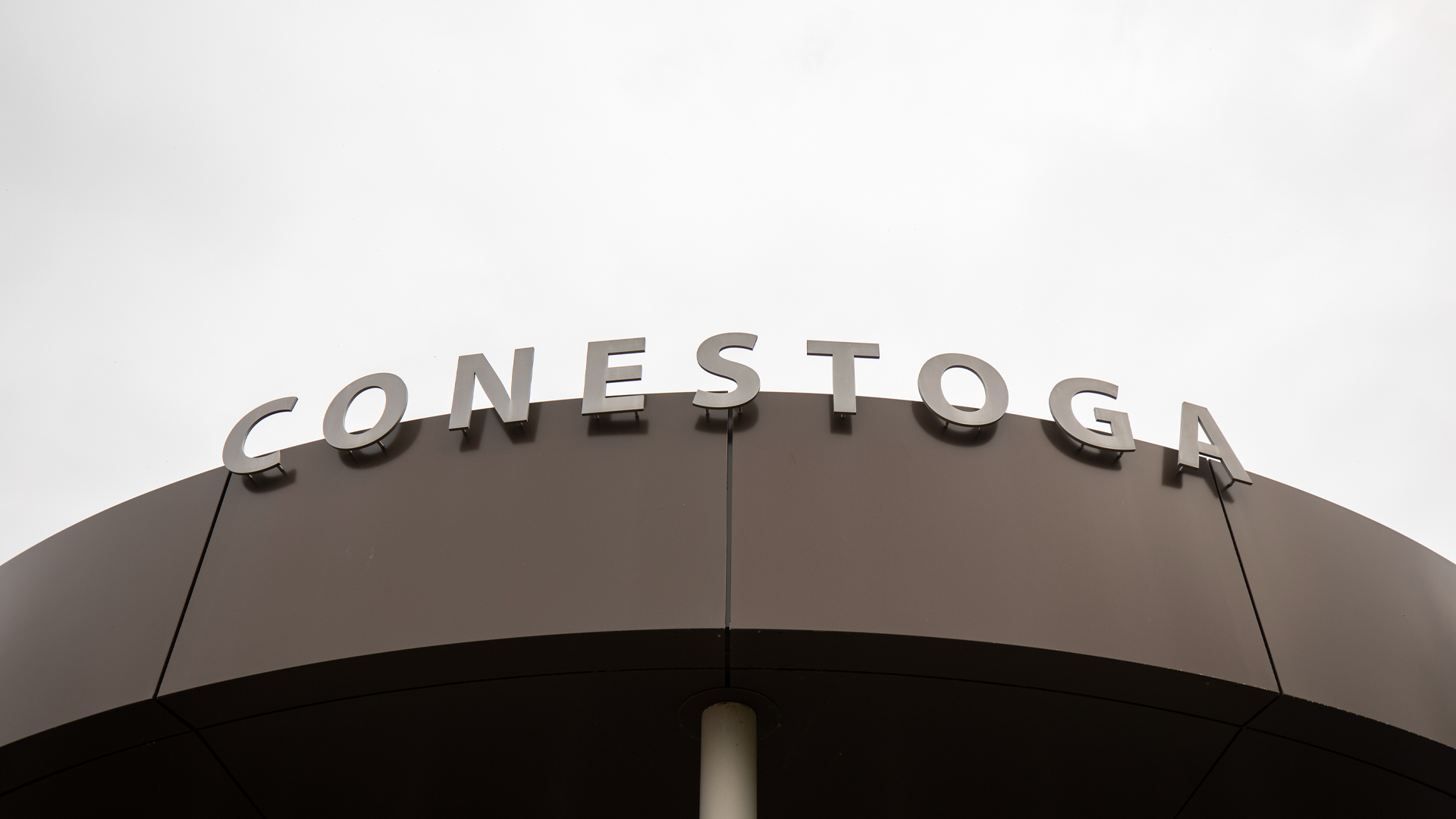 From the ground looking up, grey metal letters spell out Conestoga on a chocolate-brown piece of curved metal held up by a pole.