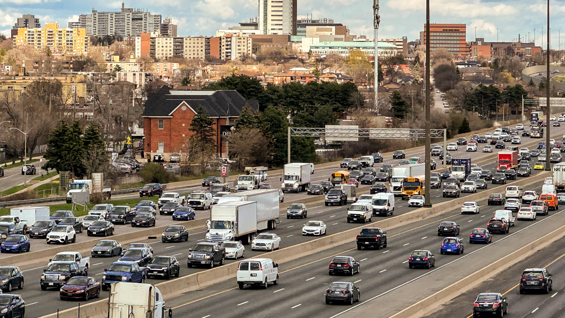 Many lanes of traffic are filled with cars. In the distance, pockets of trees are interspersed with medium high-rise towers and a mix of small apartment buildings and single-family homes.
