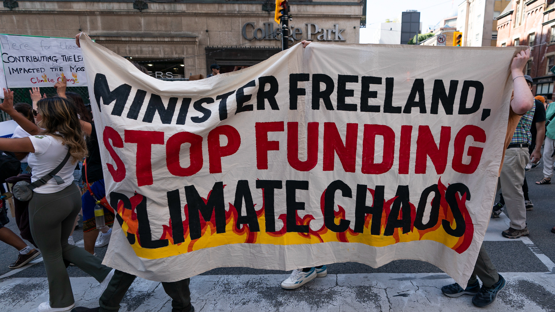 A crowd fills the street with some people holding signs. One large rectangular sheet of canvas is held up by three people. It says “Minister Freeland, stop funding climate chaos.” Yellow and orange flames are depicted as enveloping the last two words.