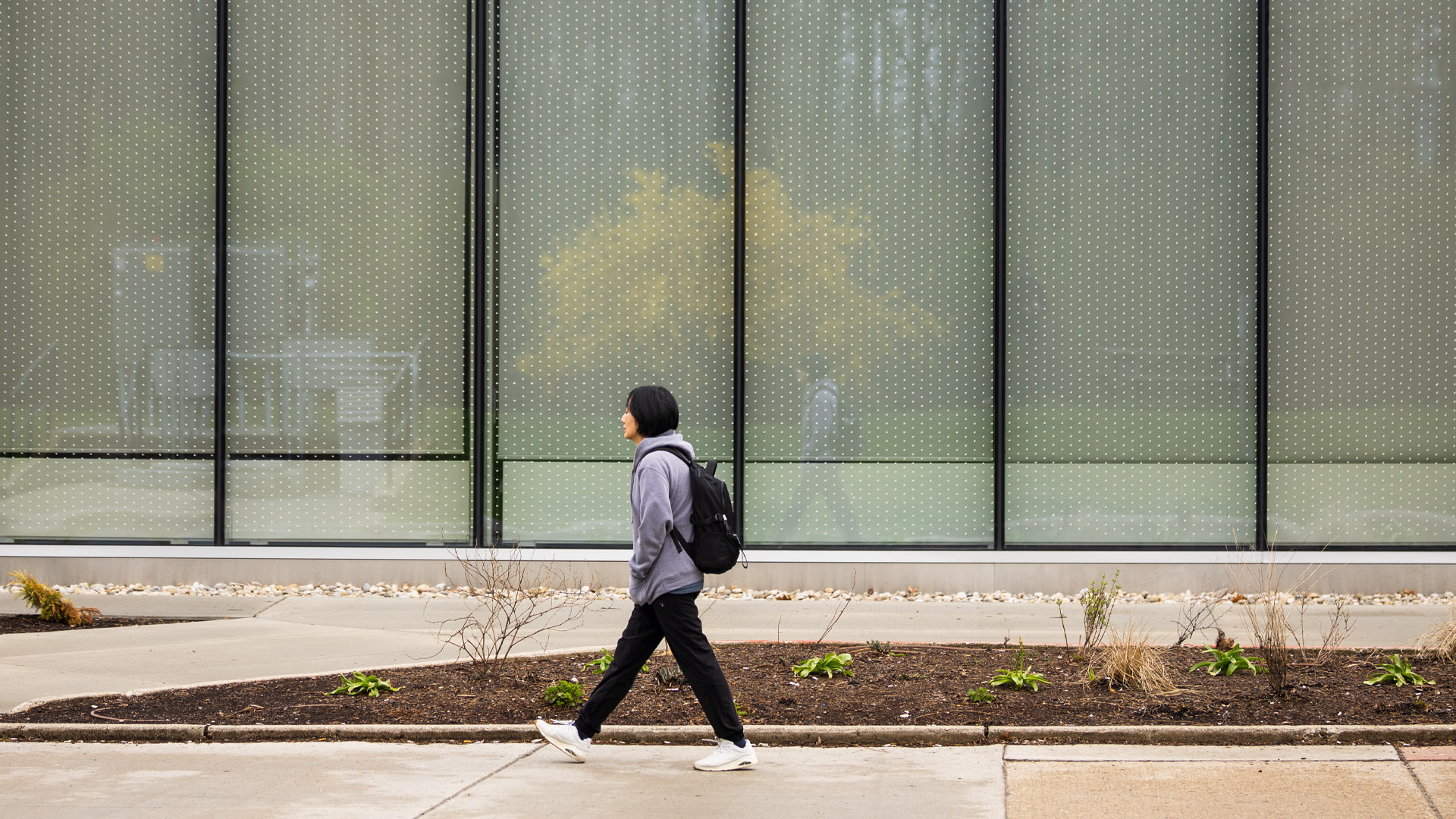 An Asian woman walks past a building with tall, rectangular floor-to-ceiling windows with black frames. She is dressed in black pants, a grey hoodie and has a black backpack.