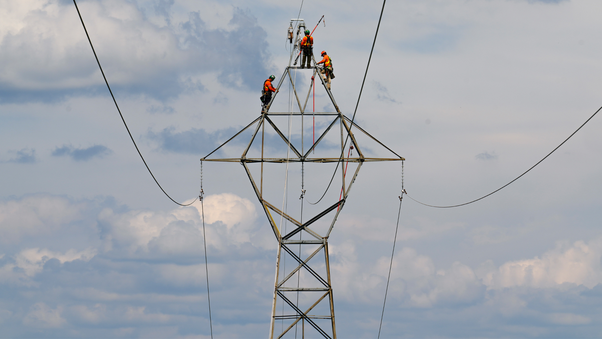 Three workers stand atop a large pylon with a cloudy sky in the background.