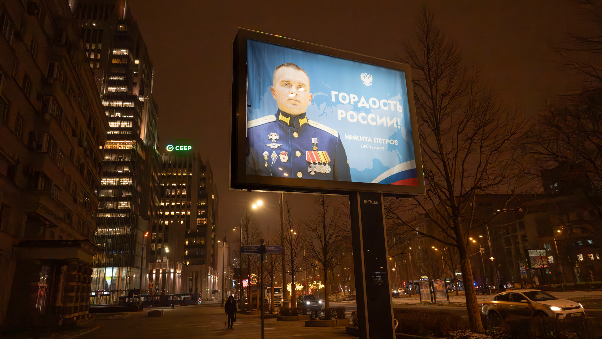 The billboard sits on a post on a sidewalk of a street with tall buildings on one side, lower buildings on the other, with leafless-trees lining both sides. It’s evening, with one person on the sidewalk. The billboard pictures a young man in a dark blue military uniform with gold epaulets and about nine medals pinned to his chest.