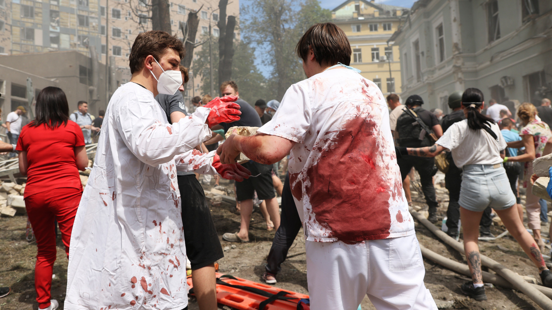 Two men in white medical coats, shirts and pants pass something not seen between them as they stand in a park next to the hospital, a two-storey building whose windows have been blown out. One man has blood spatters on his coat; the other has a large blood stain that takes up most of the back of his shirt. All around them, a crowd of people are in motion, lifting, passing, looking down. Debris covered most of the ground. Several tall trees are missing most of their trunks and branches.